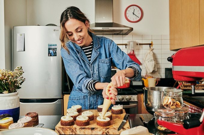 Photo of baker Lucie in an apartment kitchen putting frosting on baked goods. 