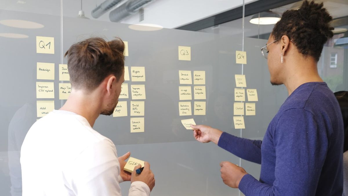 Two colleagues planning a product roadmap on a glass wall, organizing sticky-note work items by quarter.