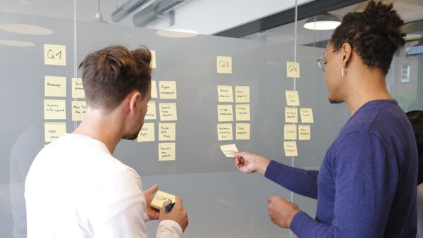 Two colleagues planning a product roadmap on a glass wall, organizing sticky-note work items by quarter.