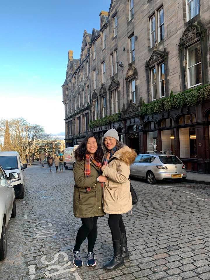 Brenda and her mum smiling together on a cobbled street in Edinburgh, bundled in coats and holding each other close on a bright winter day.