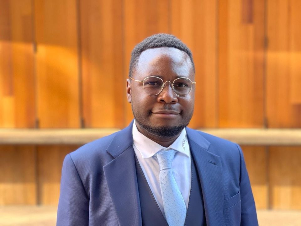 Ian Balina in a navy suit and glasses, photographed against warm architectural panels.