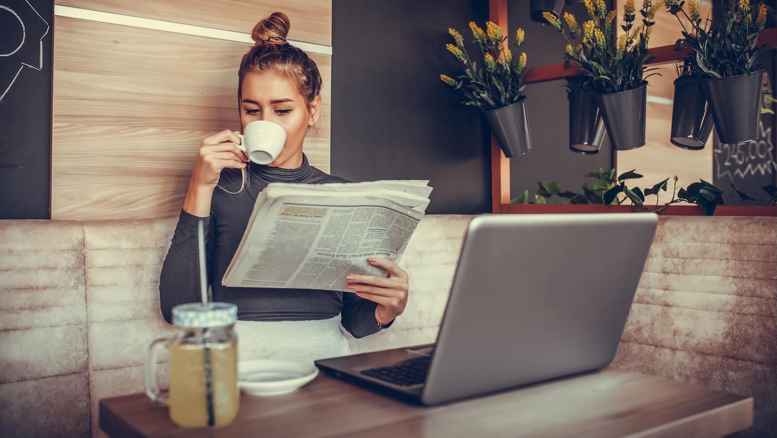 Young woman in coffee shop with laptop and newspaper drinking espresso getting ready for the market