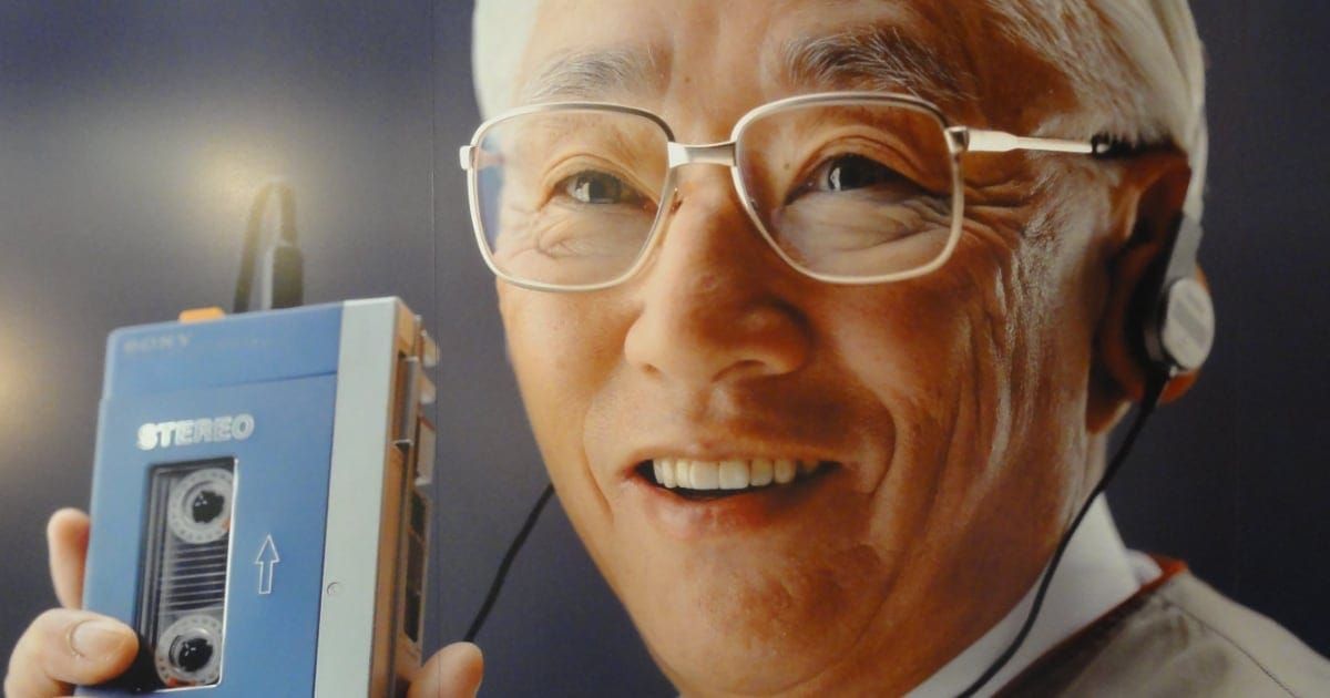 A smiling, white-haired Japanese man holds a blue Sony Walkman.