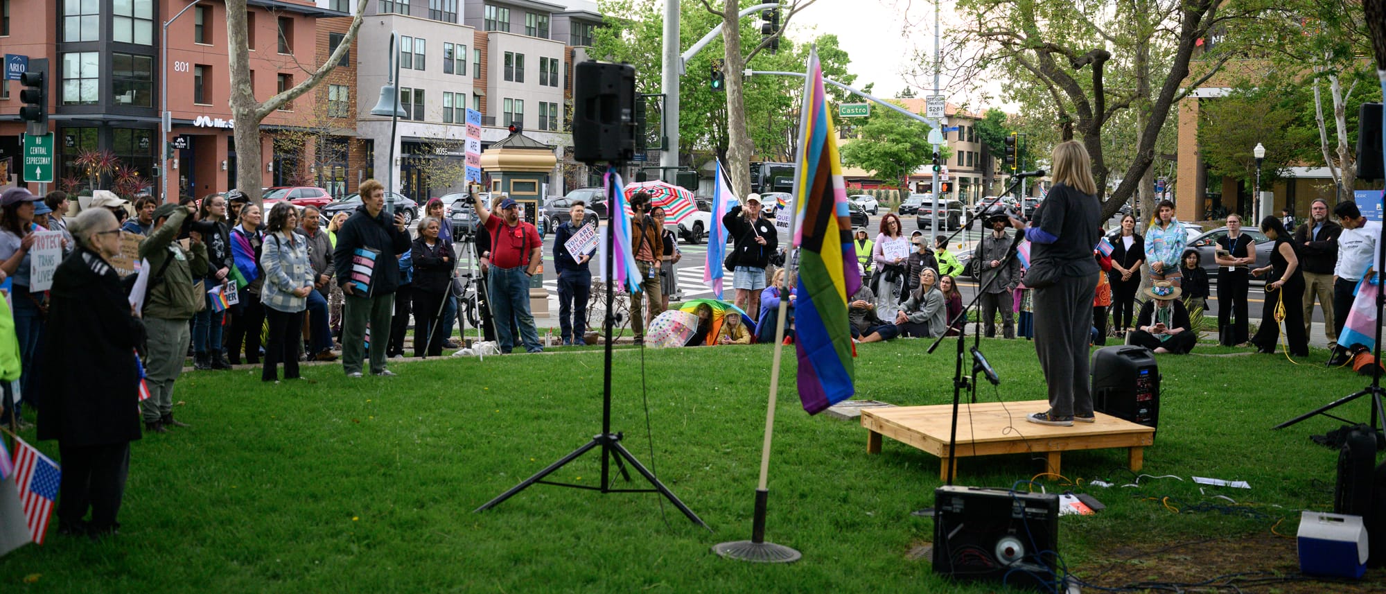 A trans woman standing on an outdoor platform speaks to a small crowd of supporters.