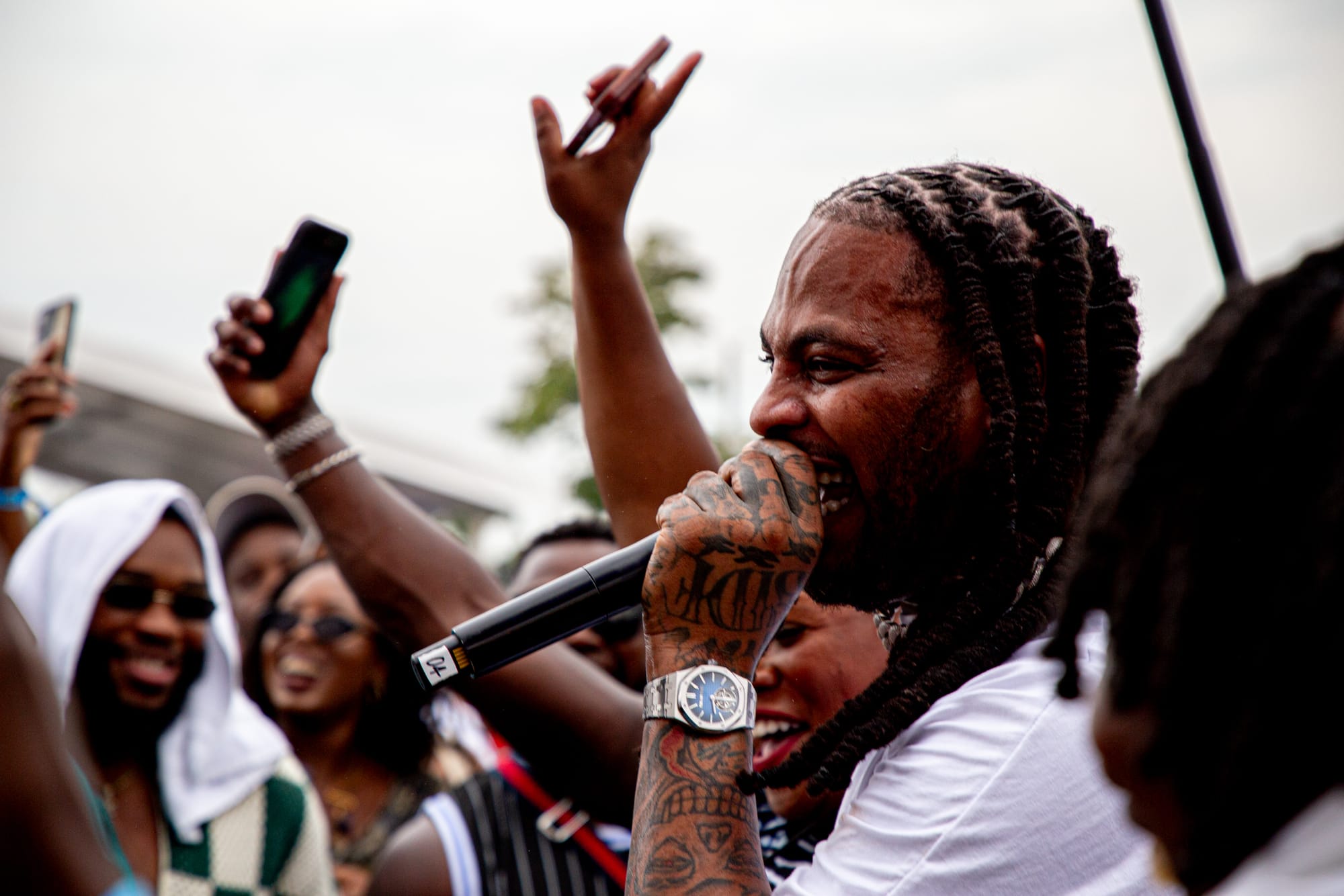 A man with braided hair and tattoos raps amid a cheering outdoor crowd as people raise their hands. 