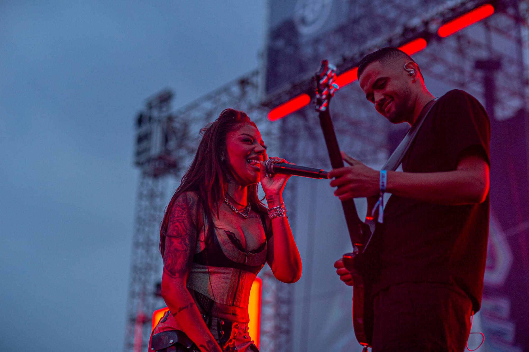 A woman sings into a microphone next to a guitarist on a stage lit in red. 