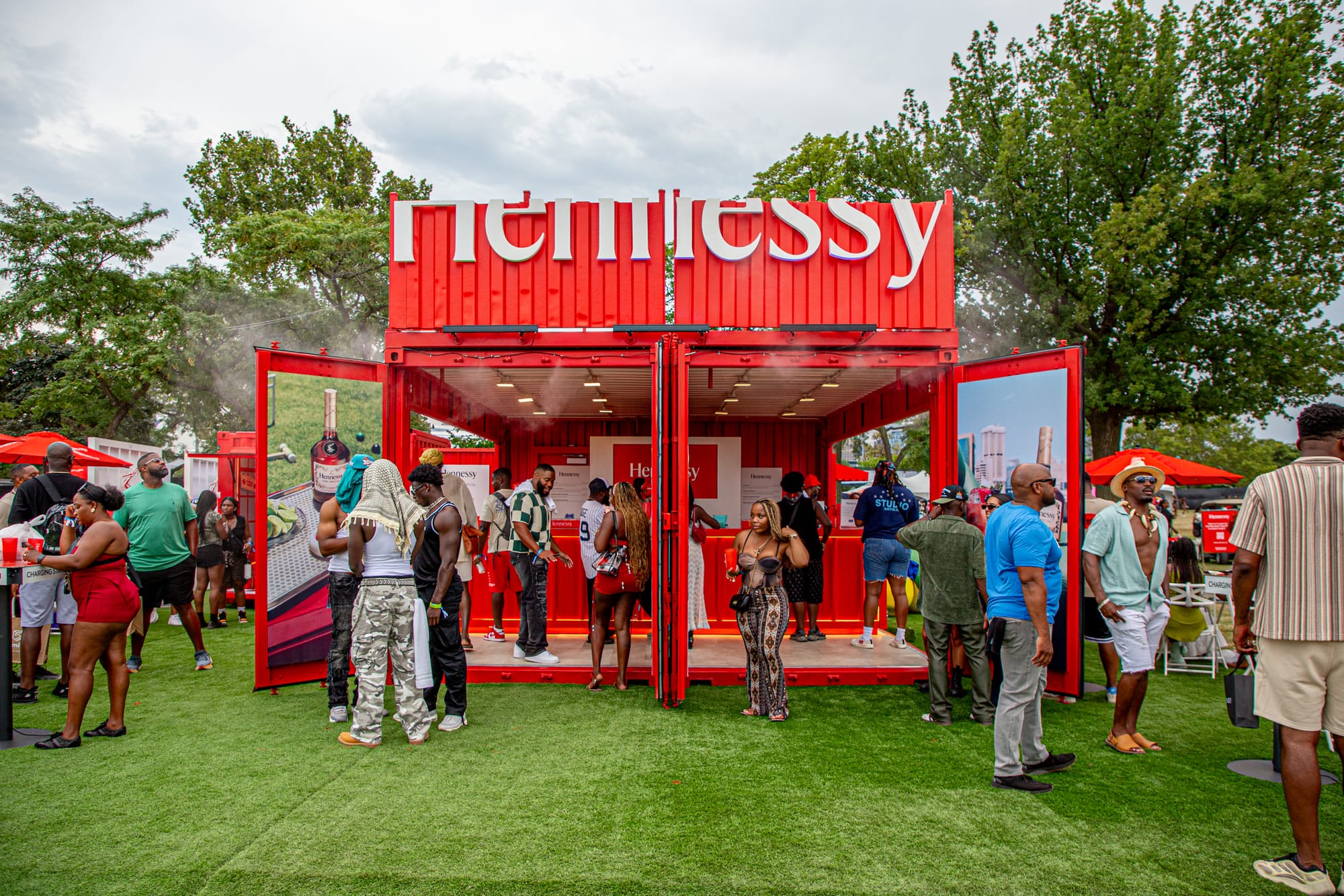 A crowd gathers at an outdoor, bright-red Hennessy-branded booth made from shipping containers on a bed of synthetic grass. 