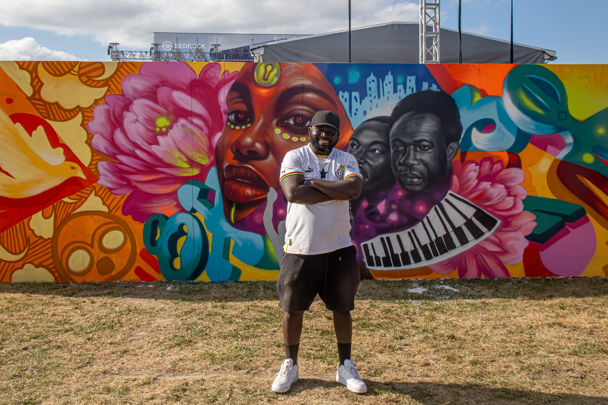 A man smiles in front of a colorful mural of three people, yellow-orange patterns, pink flowers and piano keys. 
