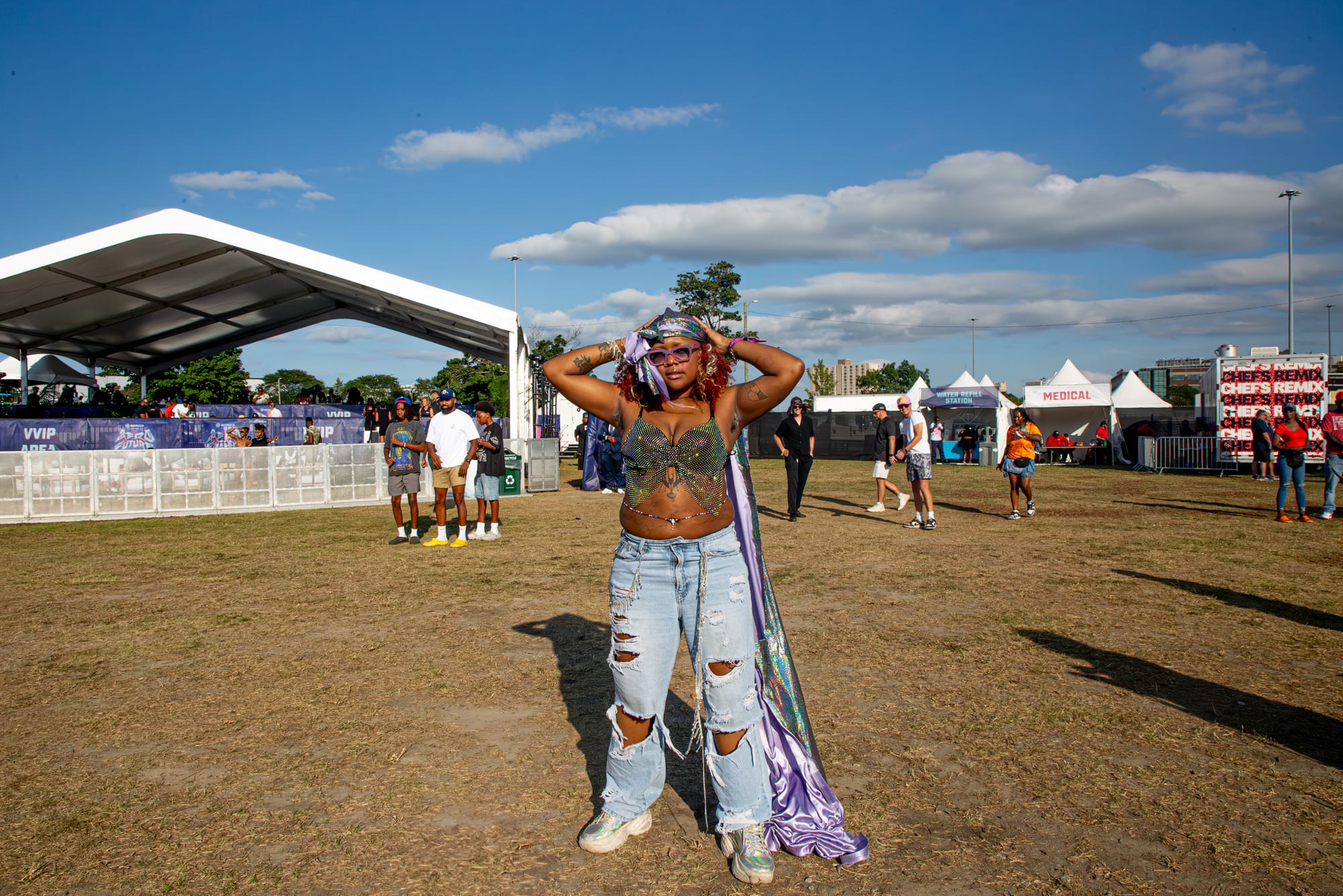 A person in ripped jeans, a beaded top and a metallic-lavender headscarf poses with arms raised at an outdoor festival. 