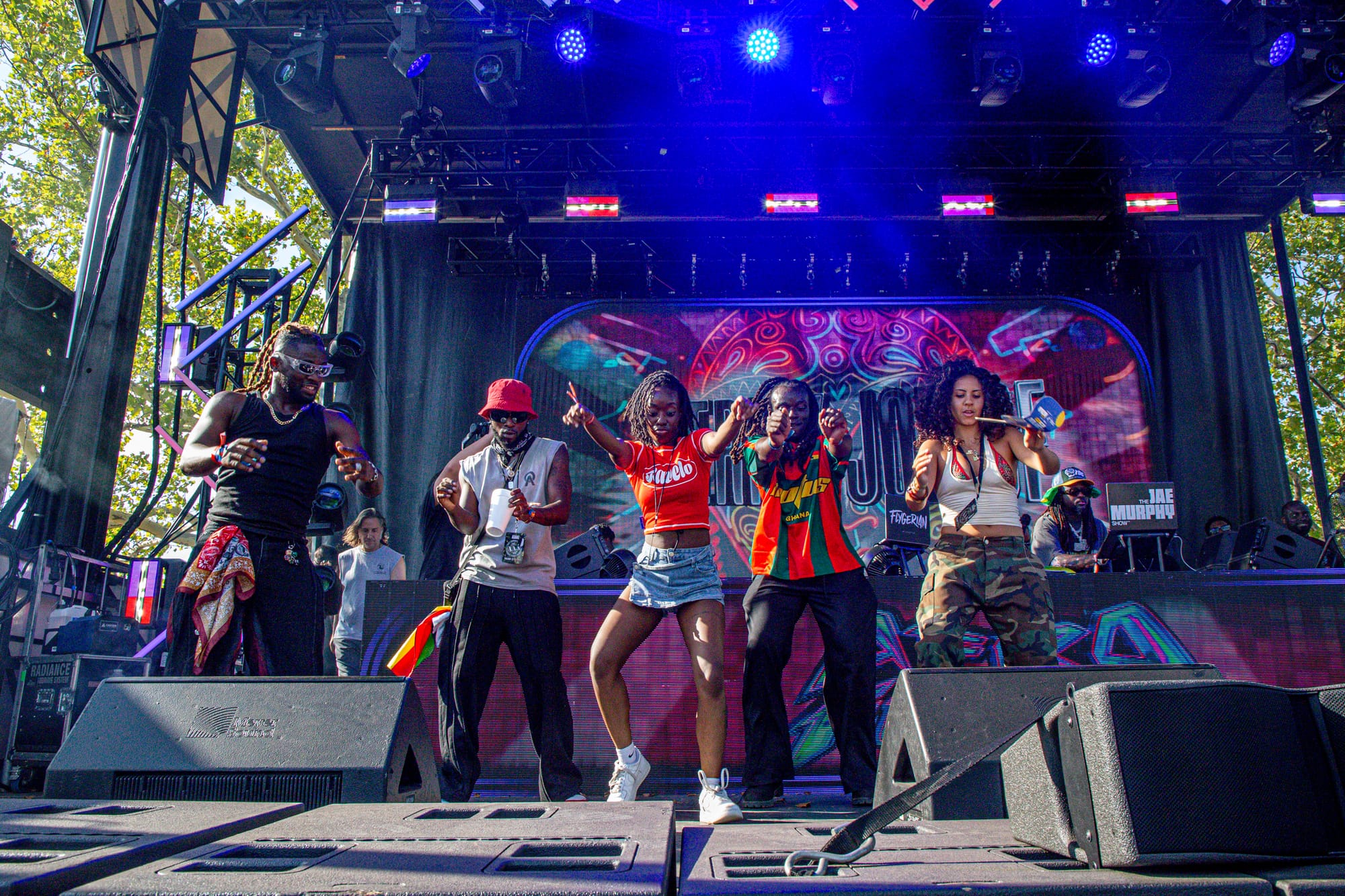 Five dancers perform on an outdoor stage with a colorful, patterned backdrop and DJ booth. 