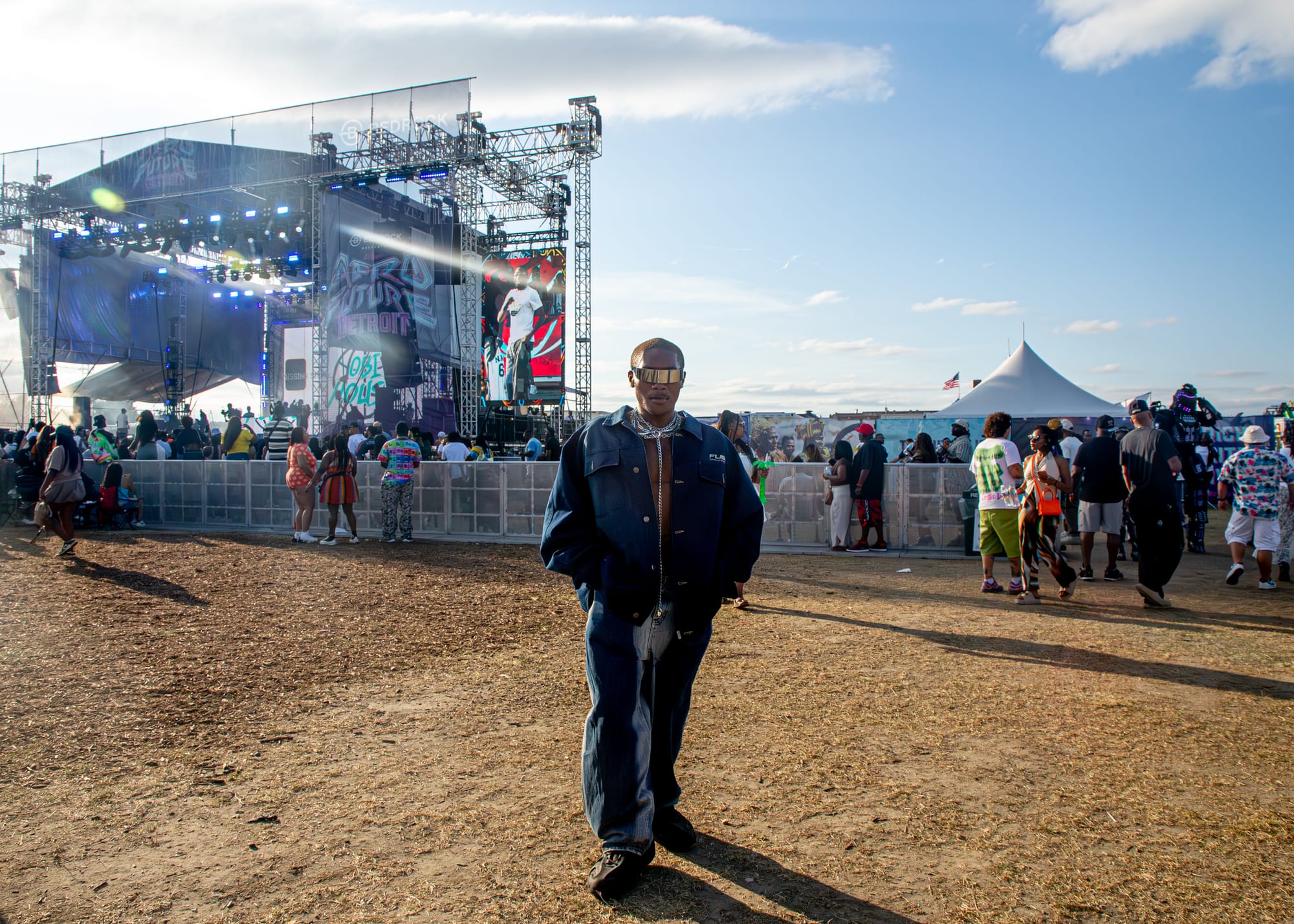 A person wearing futuristic, silver sunglasses and blue denim stands in front of an outdoor music festival stage. 