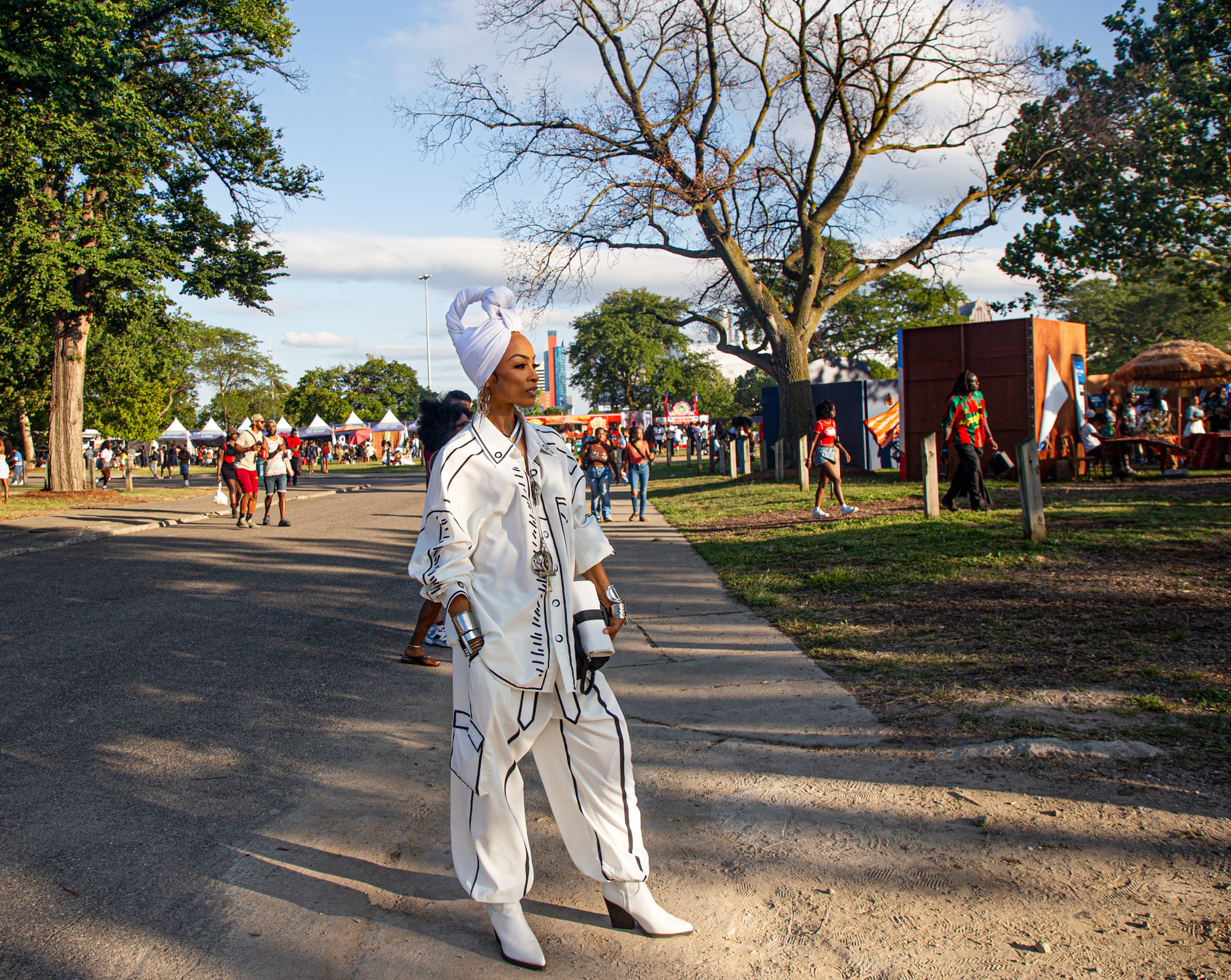 A woman in a black-and-white printed outfit and headwrap stands outdoors at a festival. 
