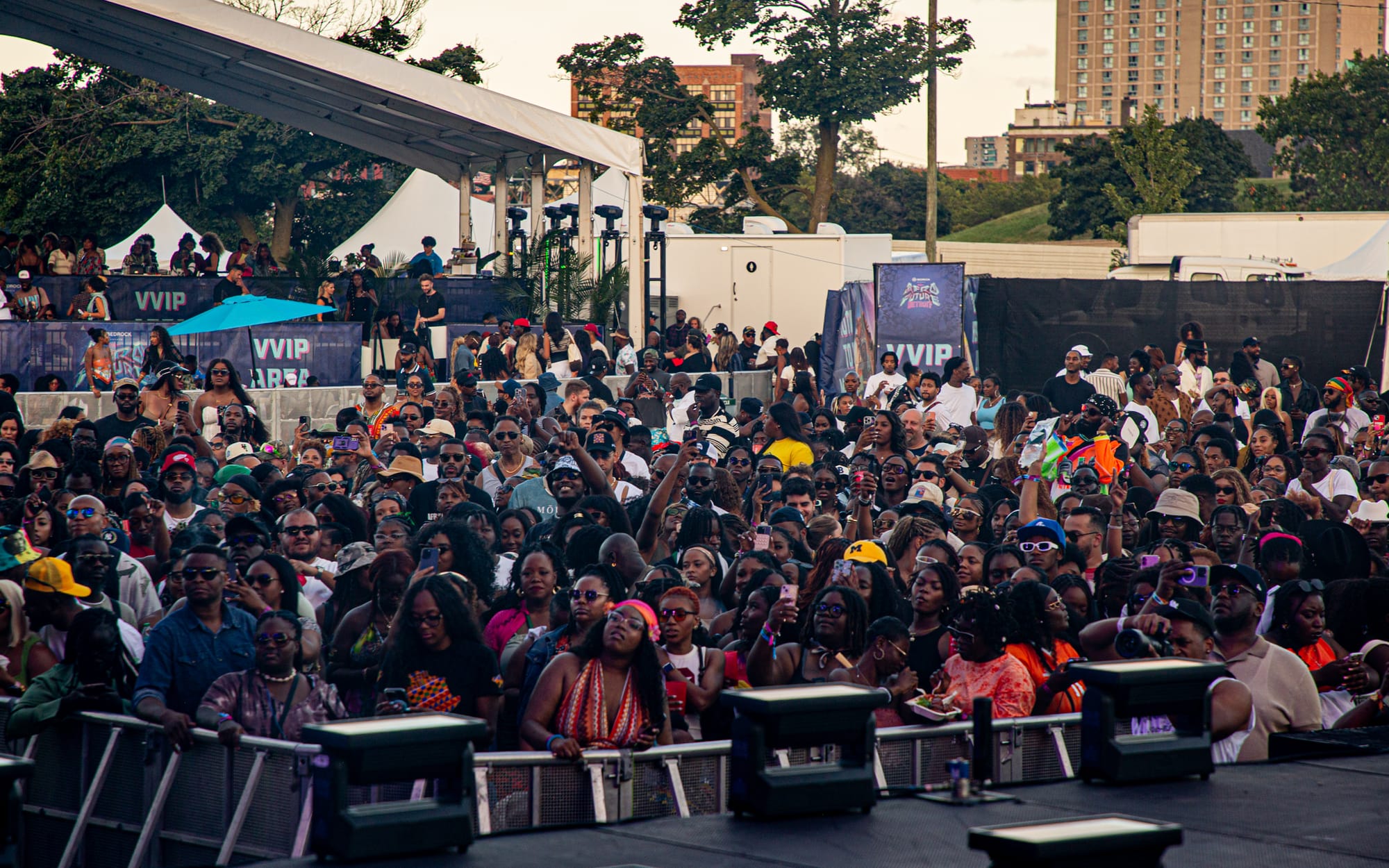 A large crowd gathers near an outdoor festival stage. Some wear sunglasses and colorful clothes. 