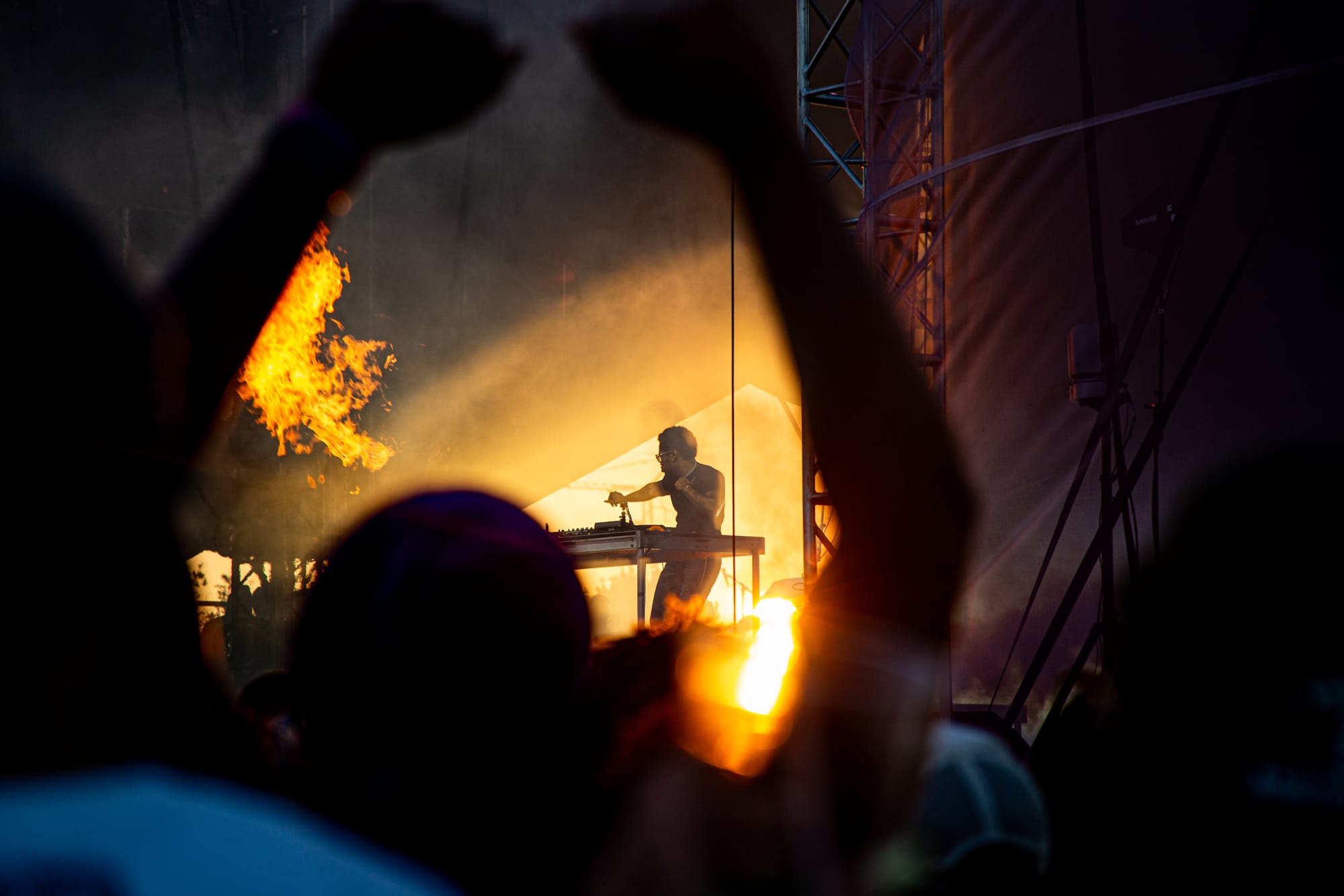 A musician performs on an outdoor, orange-lit stage with fog and flames, as silhouetted audience members raise their arms. 