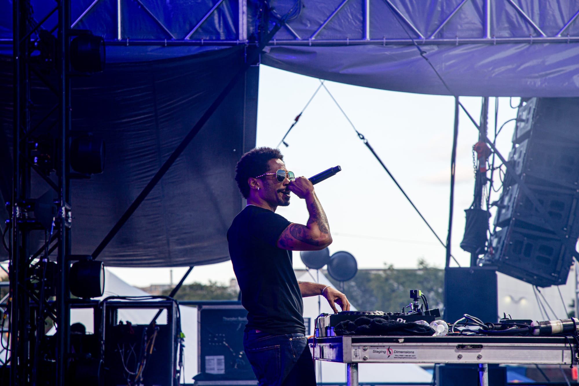 A DJ wearing sunglasses speaks into a microphone on an outdoor stage. 