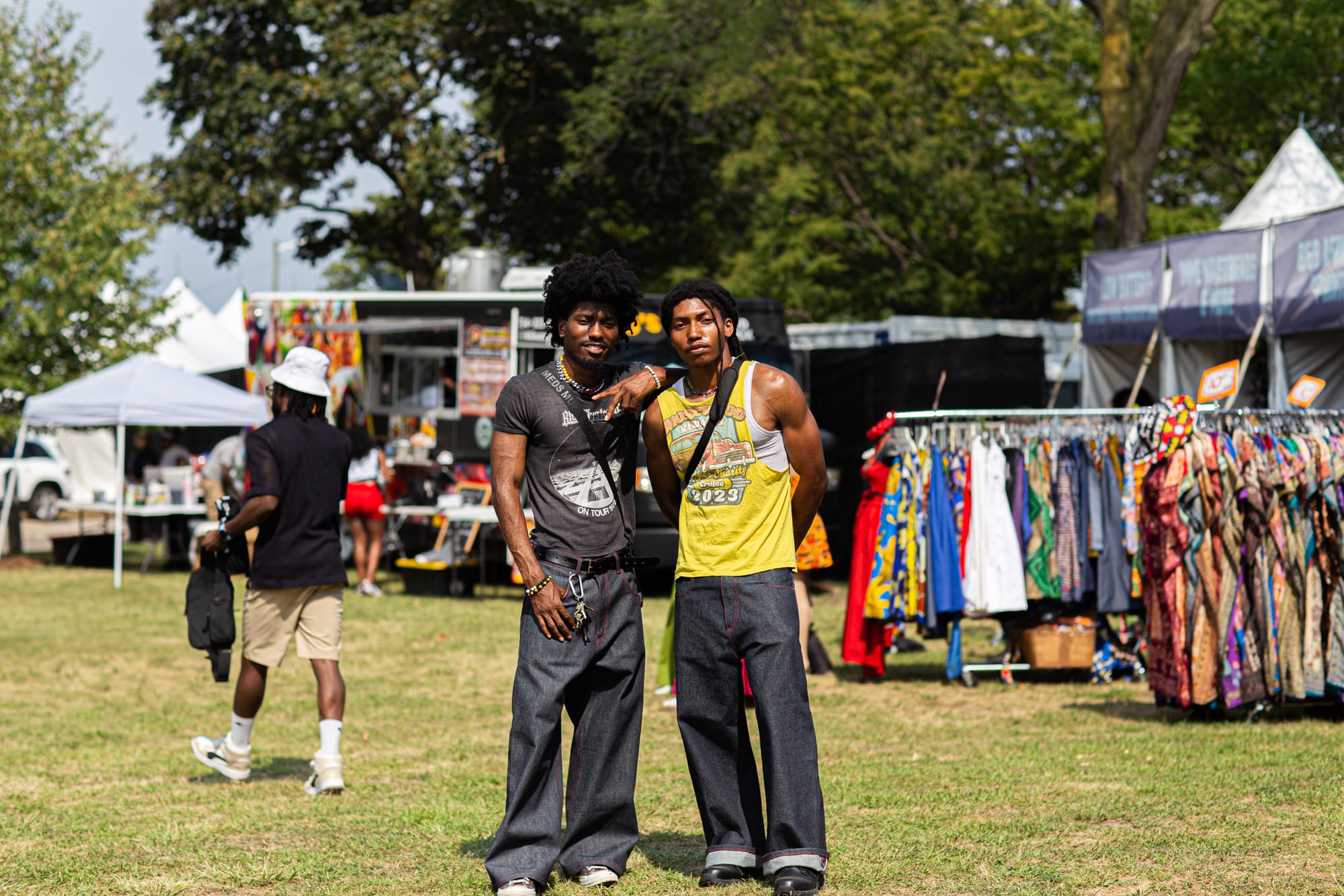Two men in black jeans and graphic tees pose together in a field with tents, clothing racks and food trucks. 