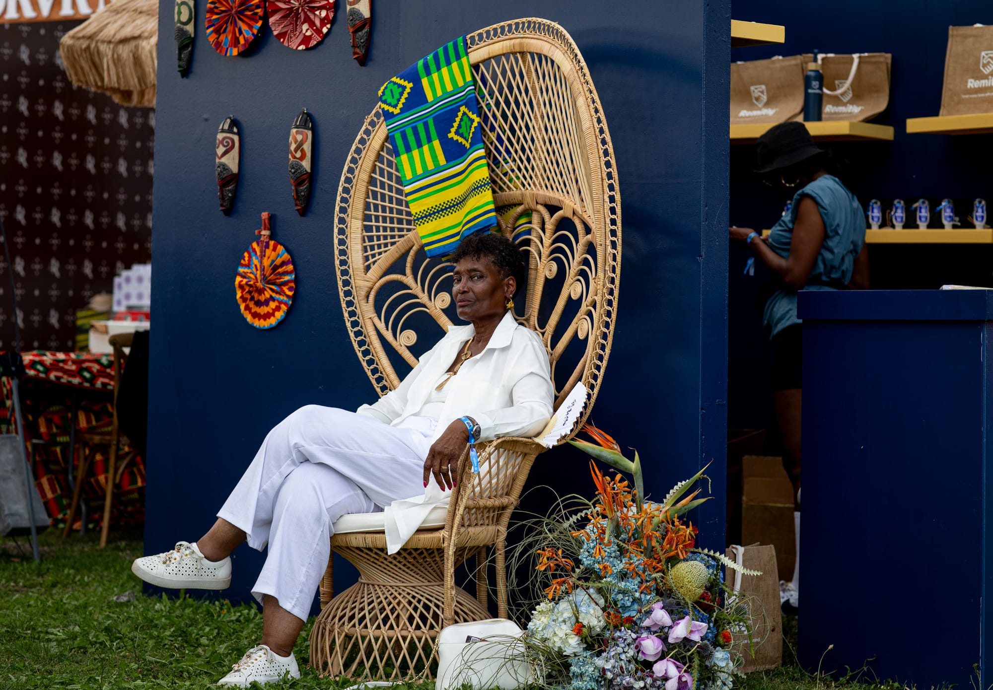 A woman dressed in white sits in a wicker peacock chair draped with a neon-green-and-blue African cloth. 