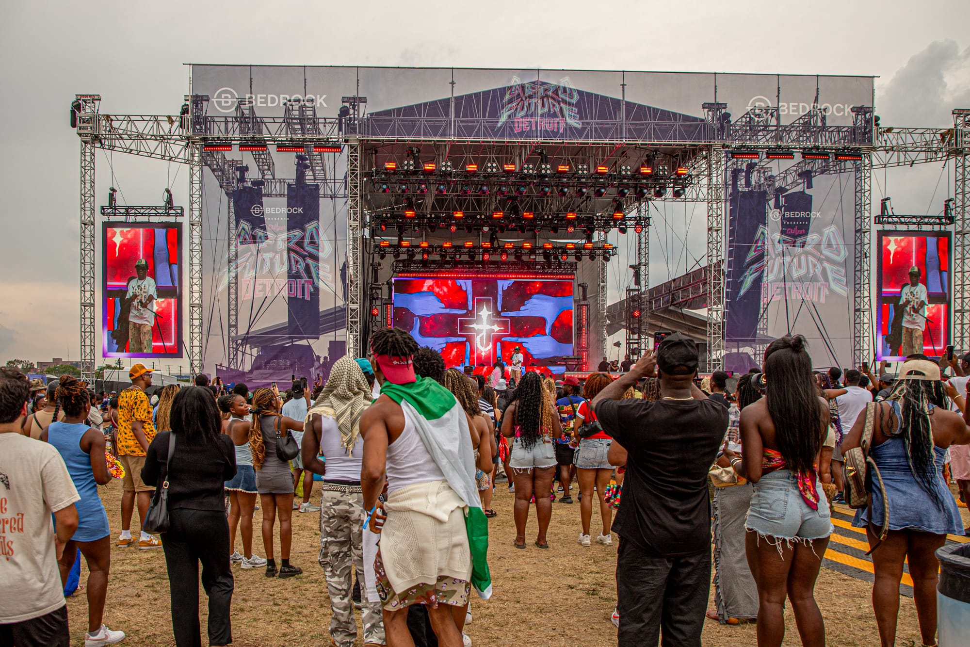A crowd watches a performer on an outdoor stage in front of a colorful animation on a screen. 