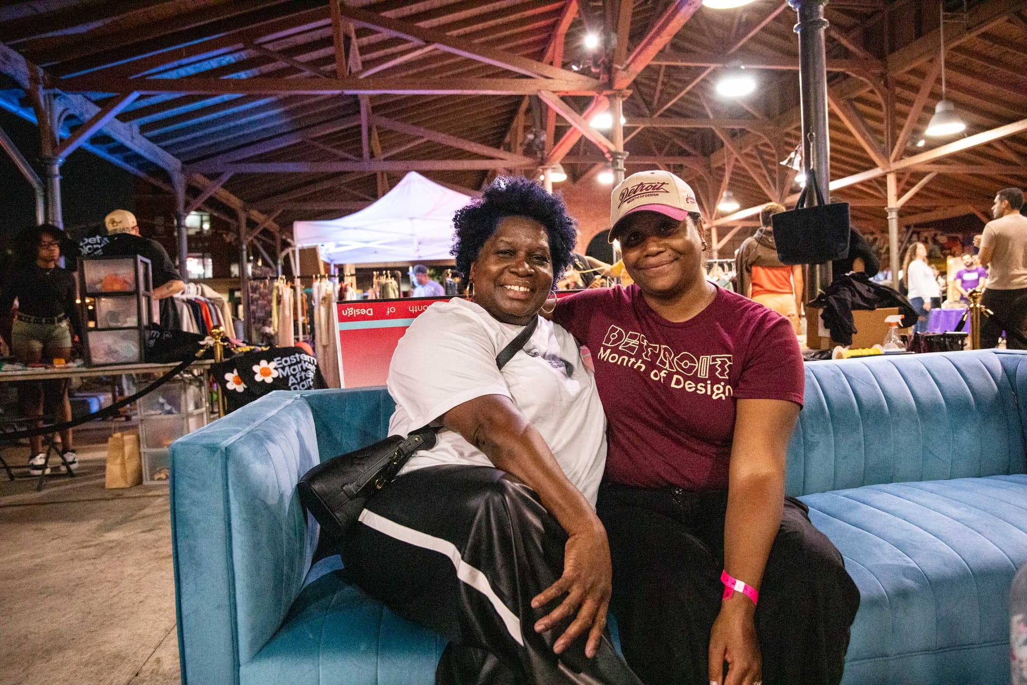 Two women sit together smiling on a blue couch at an indoor market.
