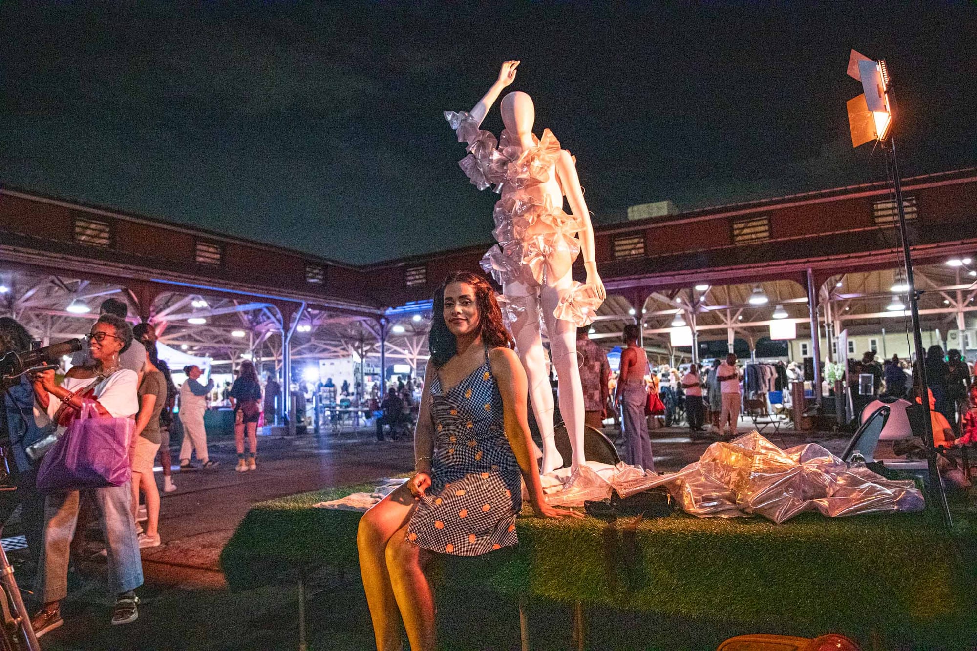 A woman in a polka-dot dress sits on a table outdoors at night, with a white mannequin dressed in ruffled fabric behind her.