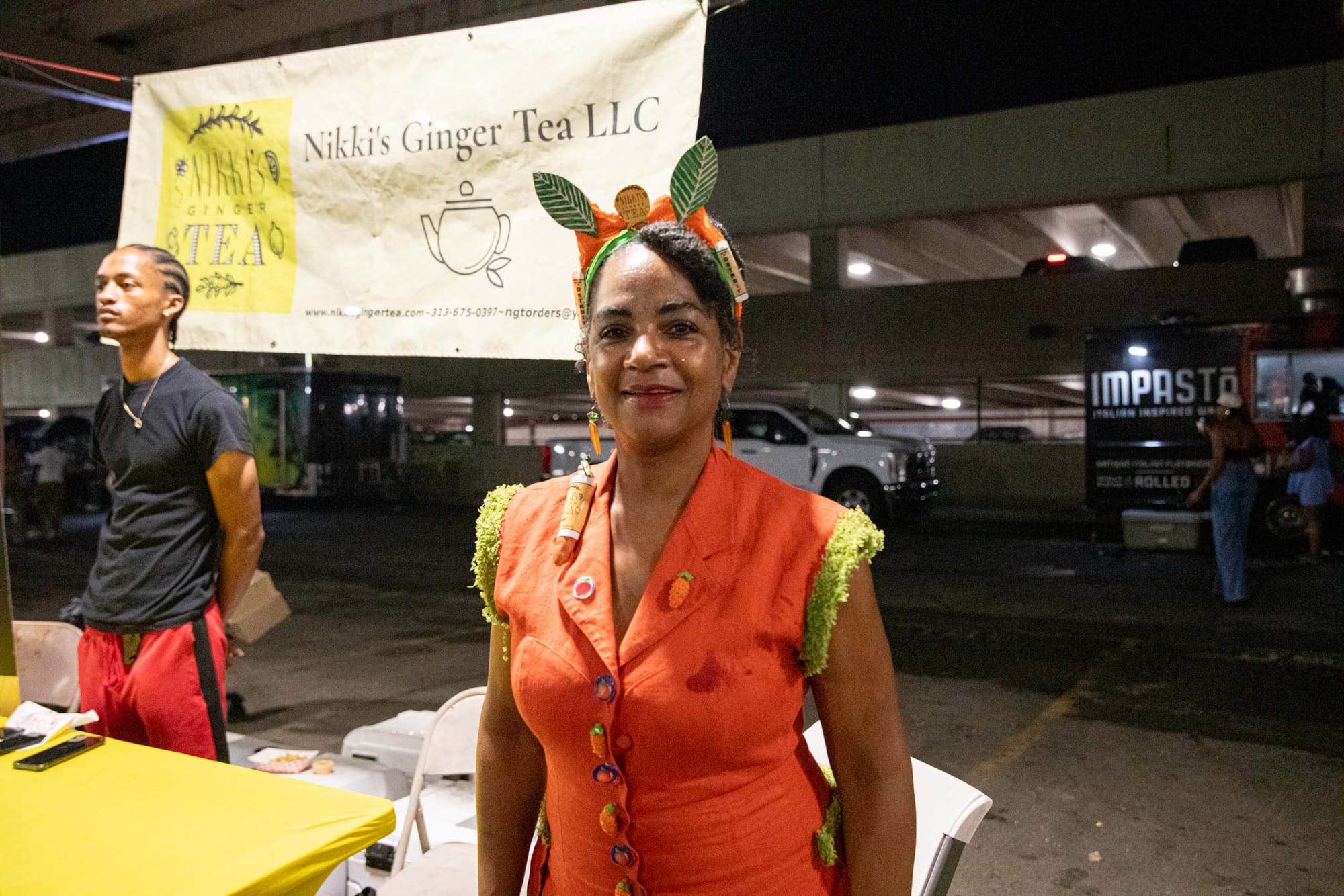 A woman in an orange dress and leafy headband smiles at a market stall for Nikki’s Ginger Tea LLC.