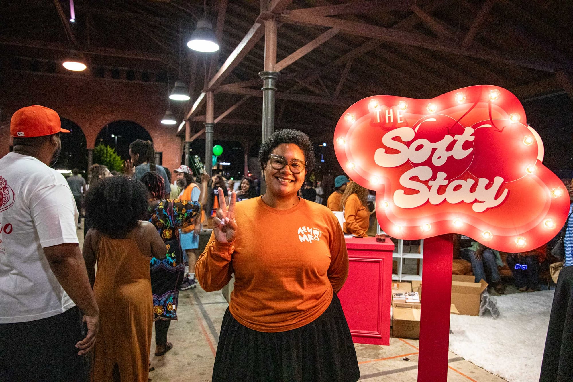 A woman wearing an orange shirt smiles with the peace sign up. Behind her is a glowing sign that reads “The Soft Stay”