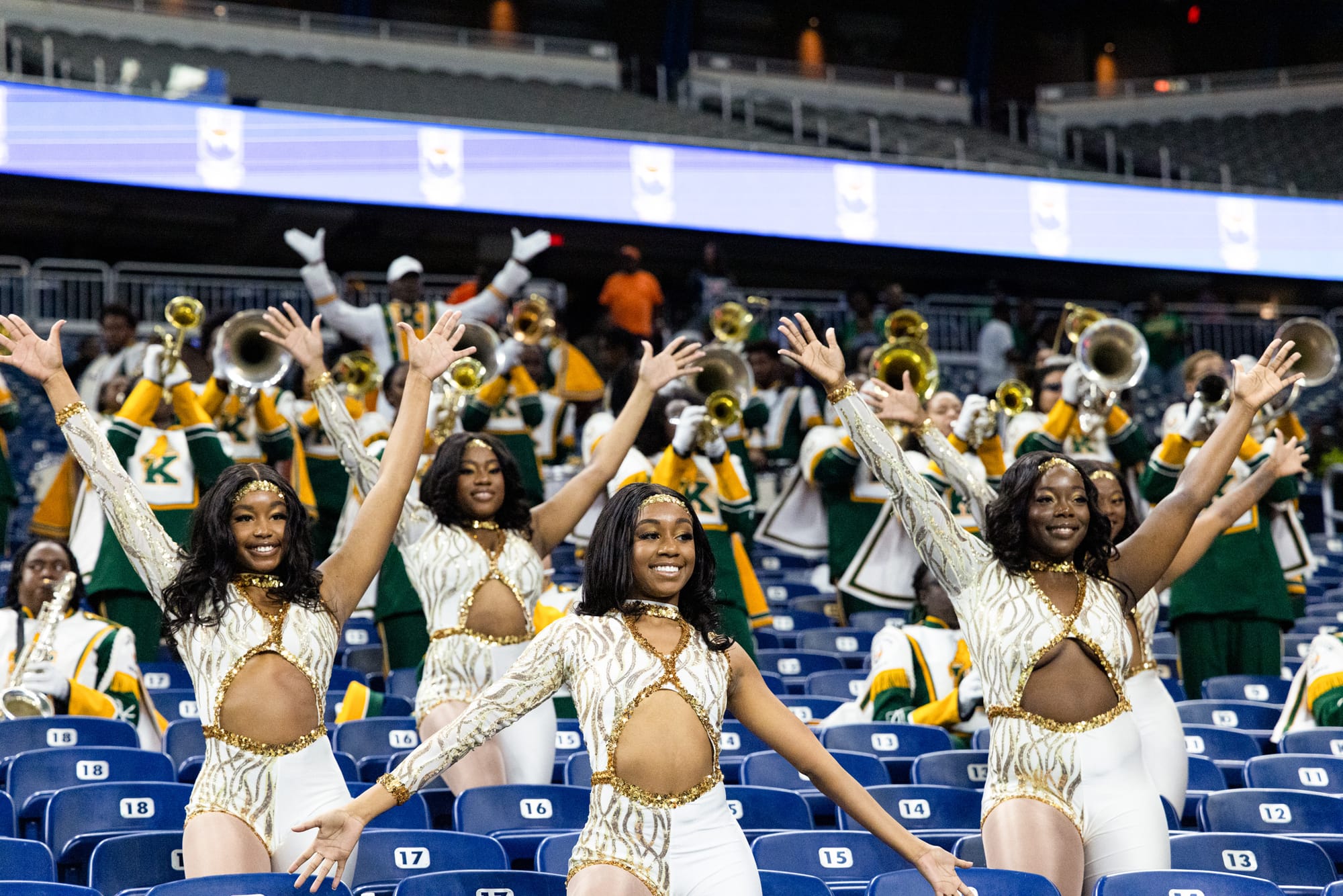 Five dancers in white and gold costumes perform with their arms raised in front of a marching band in a stadium with blue seats.