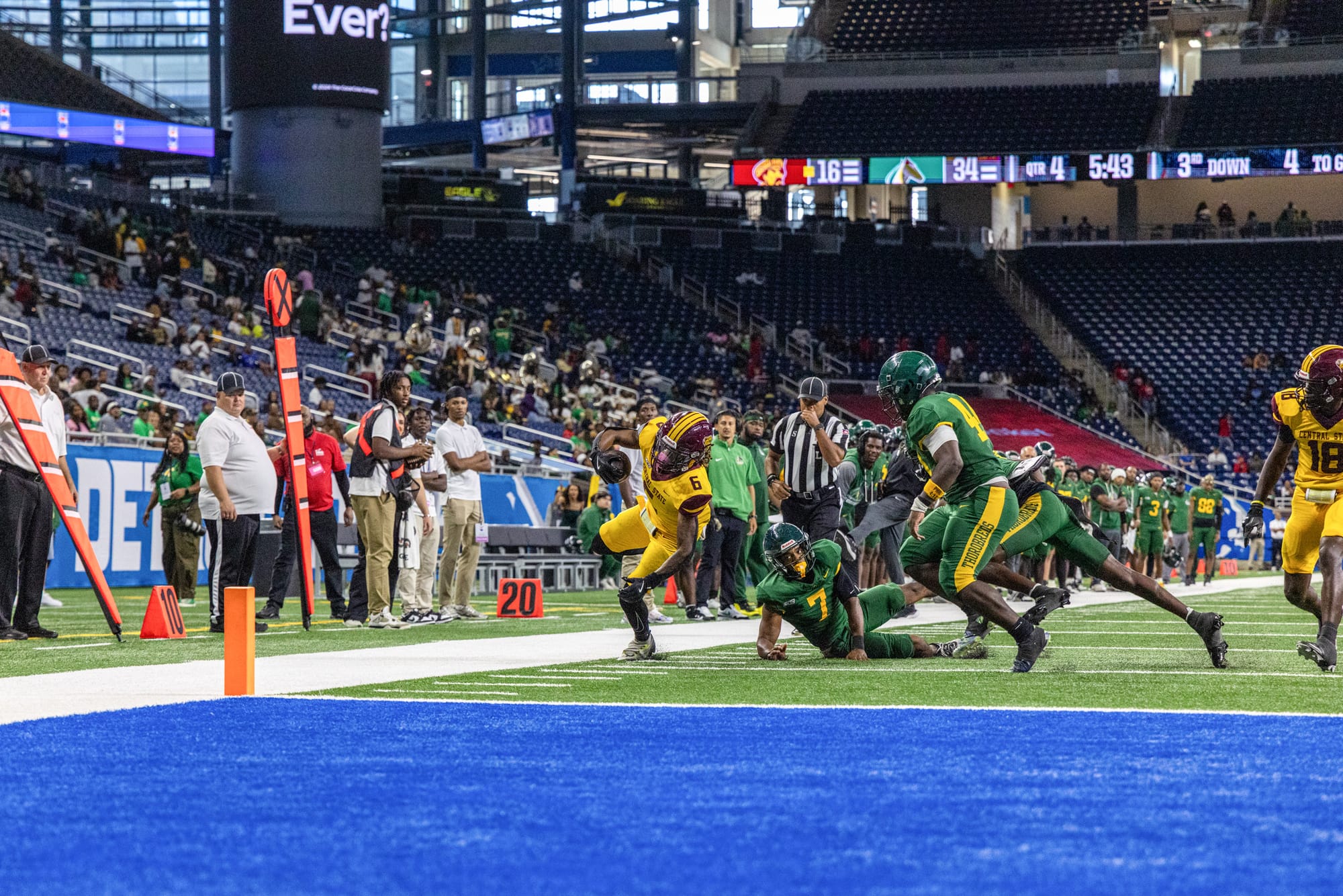 A football player in yellow dives toward the end zone while two defenders in green attempt to tackle him near the sideline with officials nearby.