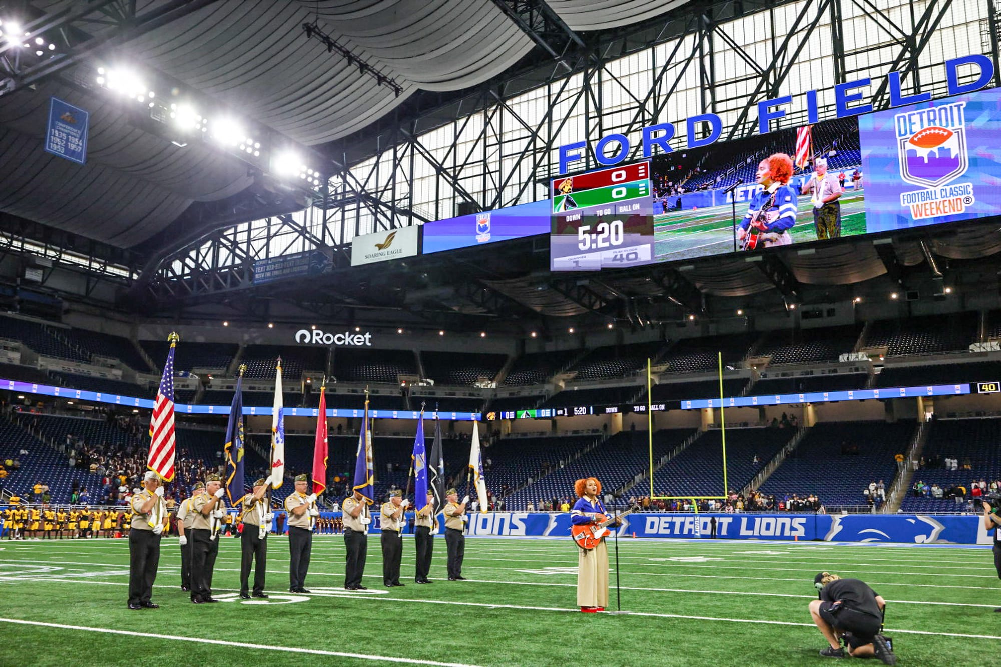 A color guard stands on a field holding flags, while a person with orange hair performs on the field of an indoor football stadium.