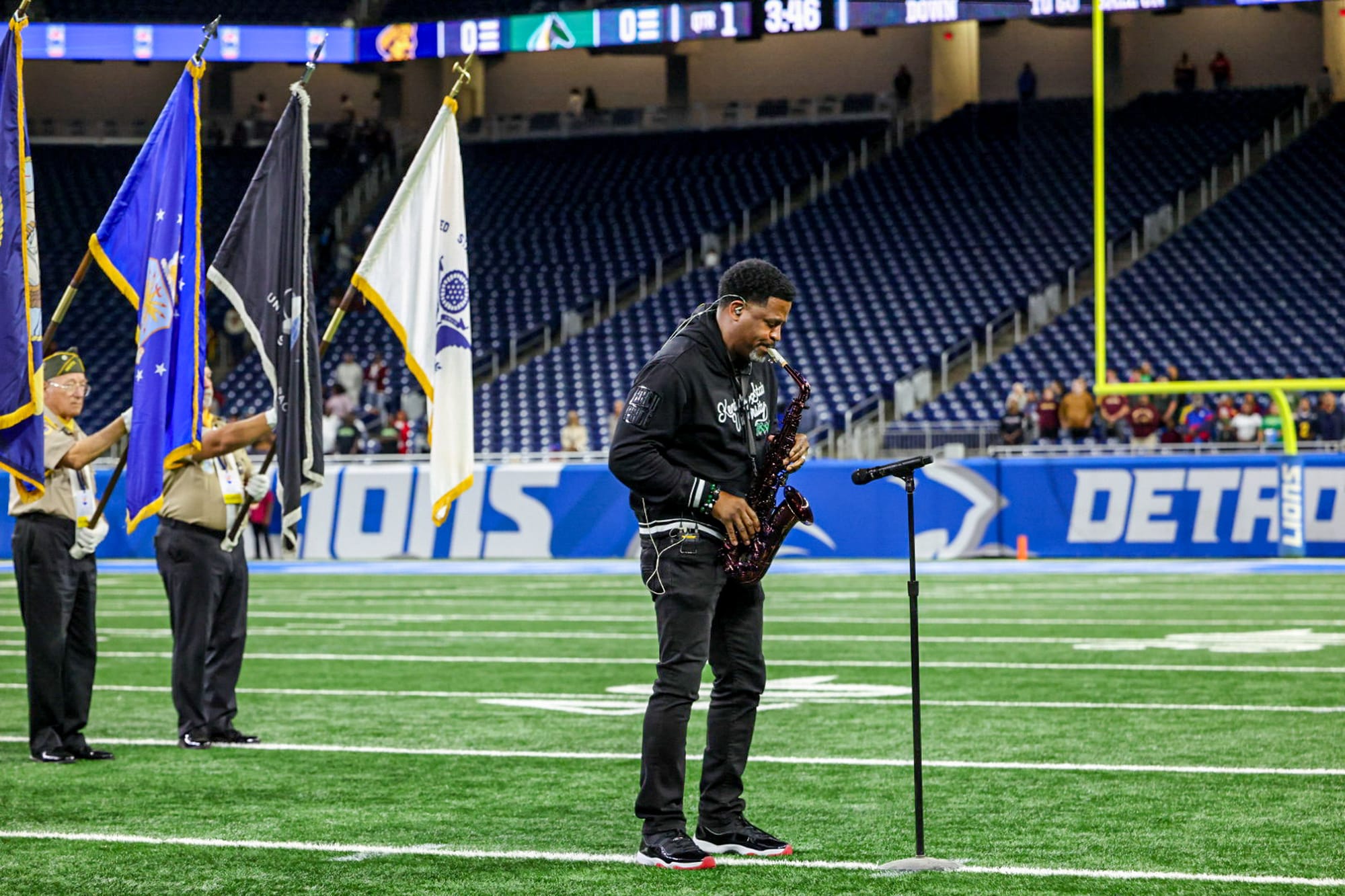 A musician plays the saxophone on an indoor football field near a microphone, with color guards holding flags in the background.