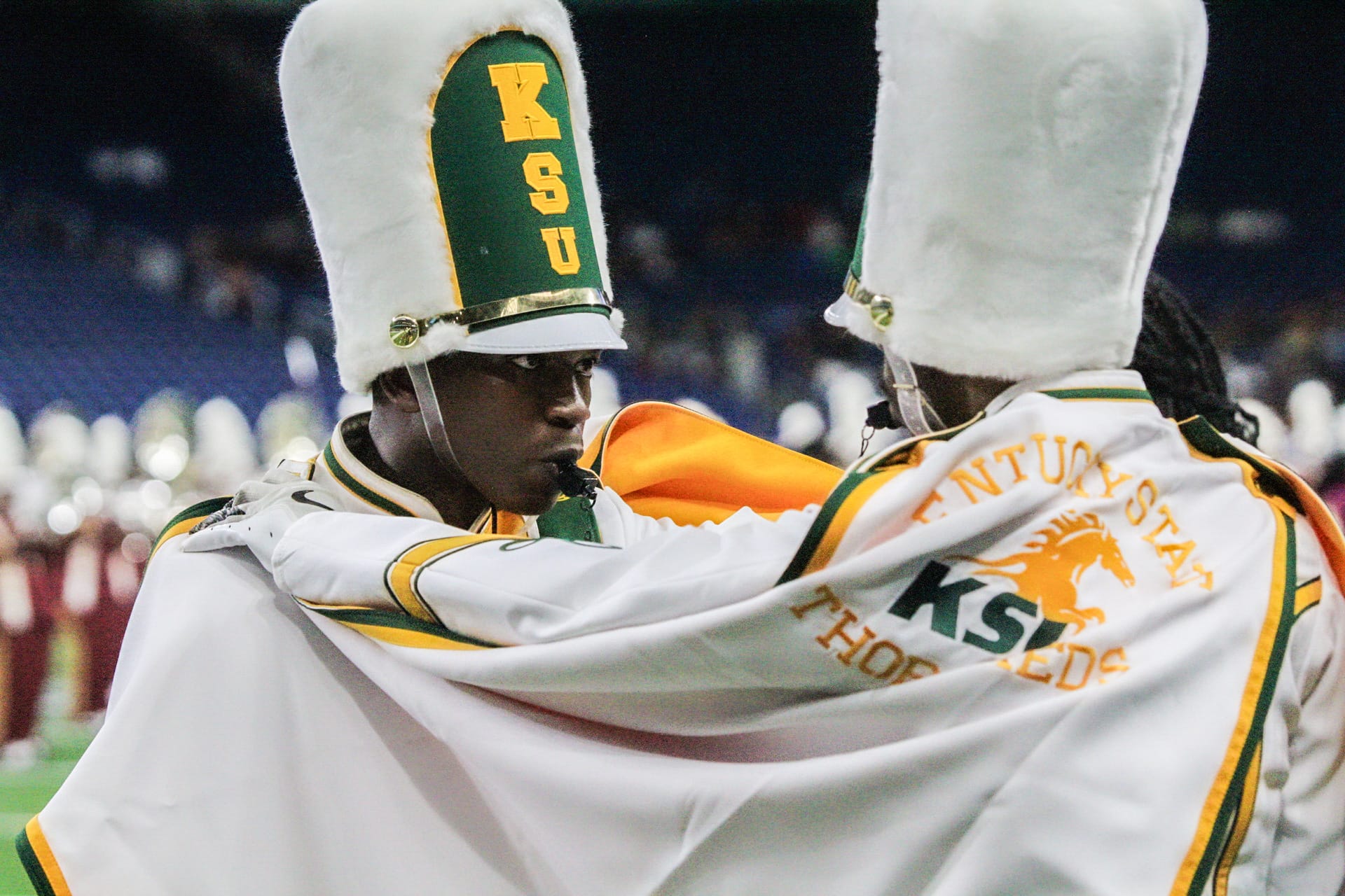 Two marching band members in tall hats and uniforms stand face to face, one with a whistle, while adjusting each other's capes.