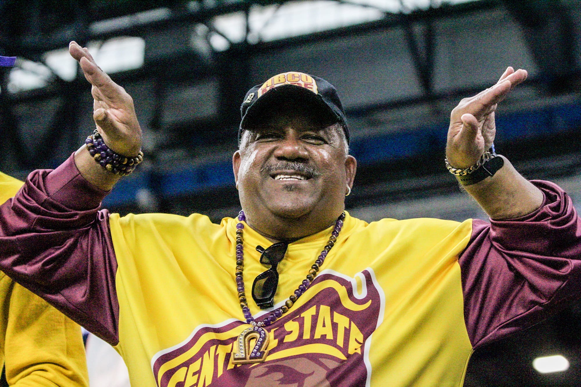 A smiling man in a yellow and maroon Central State shirt and hat, with purple and gold beaded jewelry raises his arms.