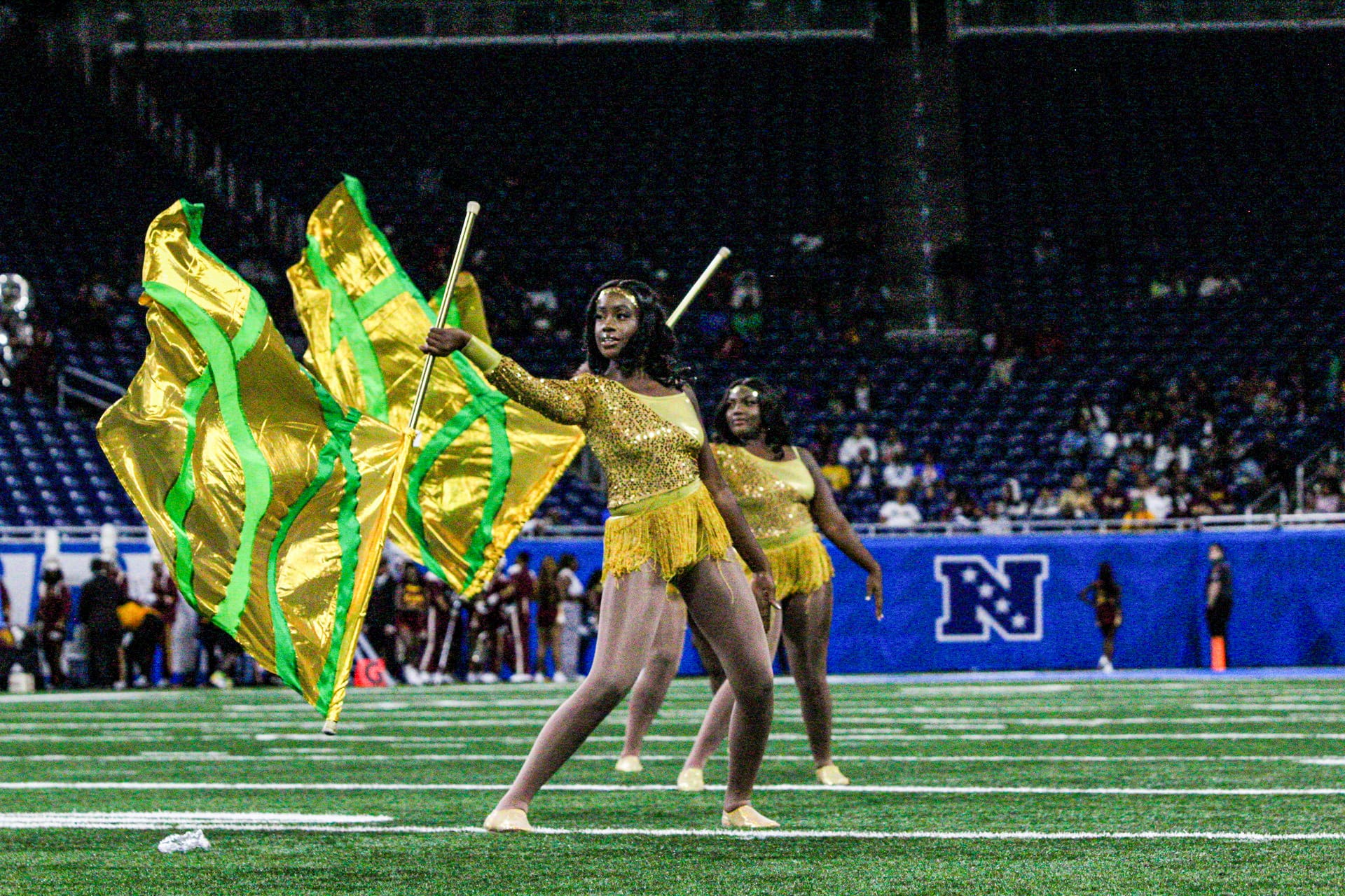 Two dancers in gold costumes perform a flag routine on an indoor football field, each holding large yellow and green flags.