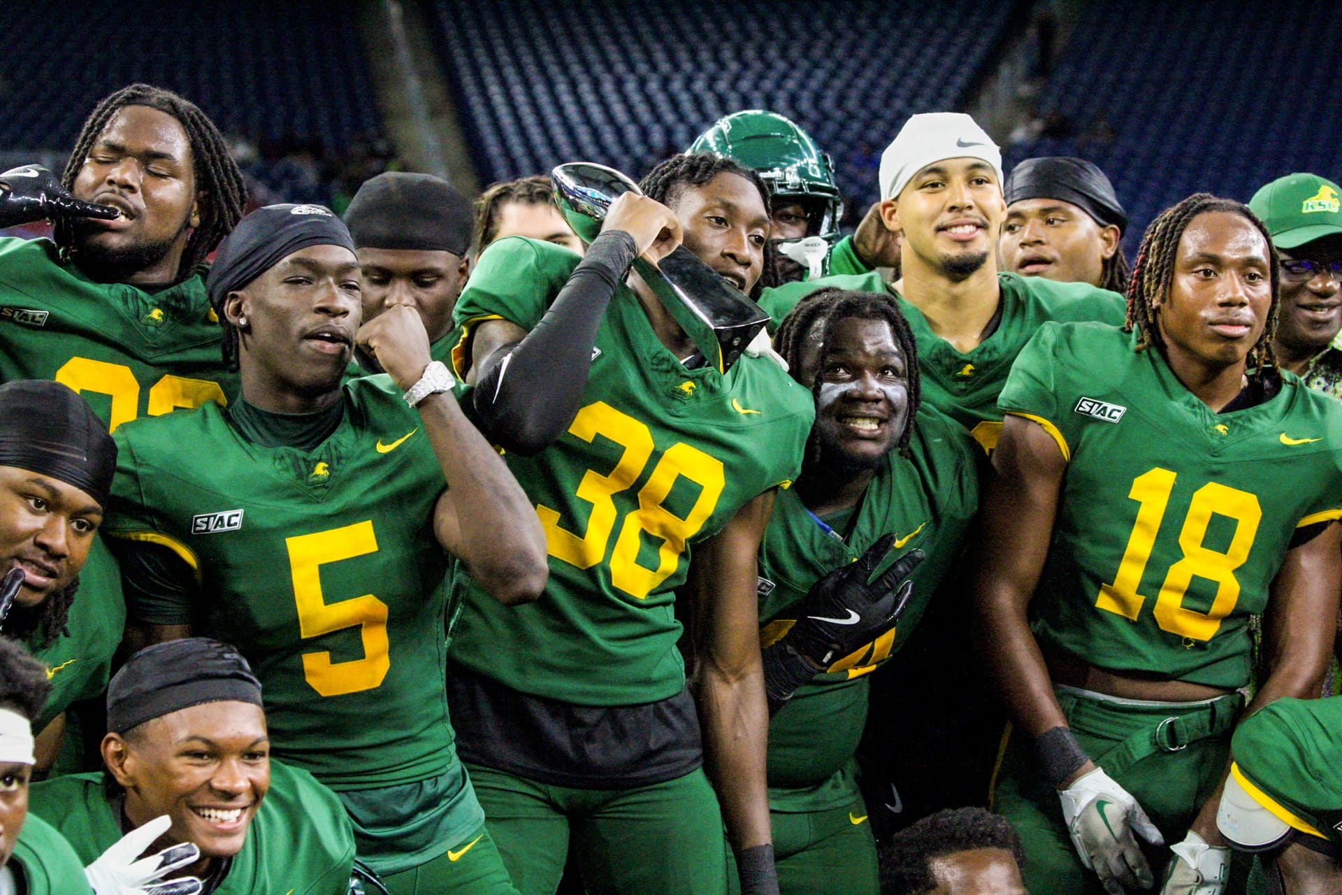A group of football players in green and yellow uniforms pose together while one holds a trophy.