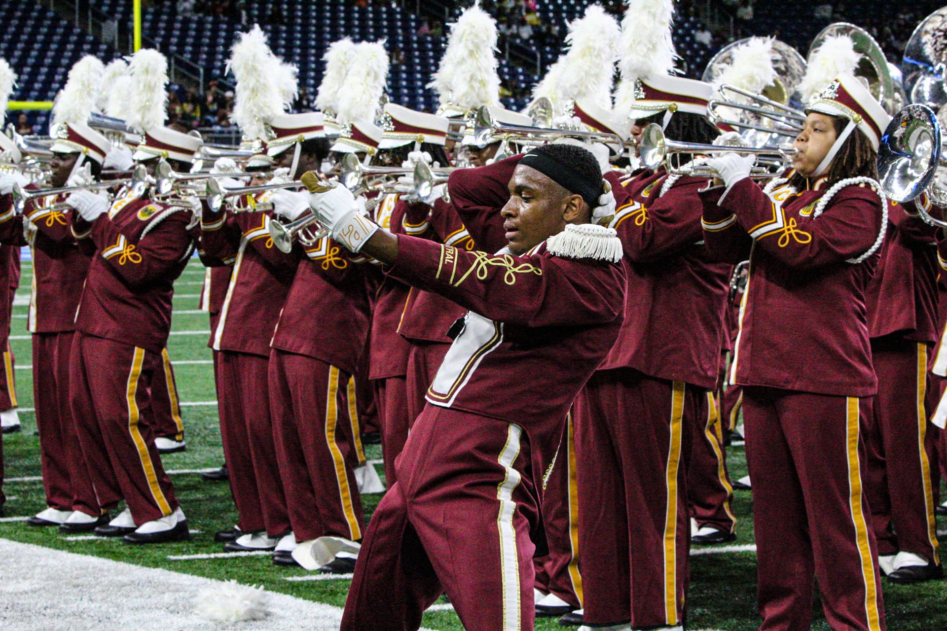 A marching band in maroon and gold uniforms performs on a football field, with one member in front posing while others play brass instruments.