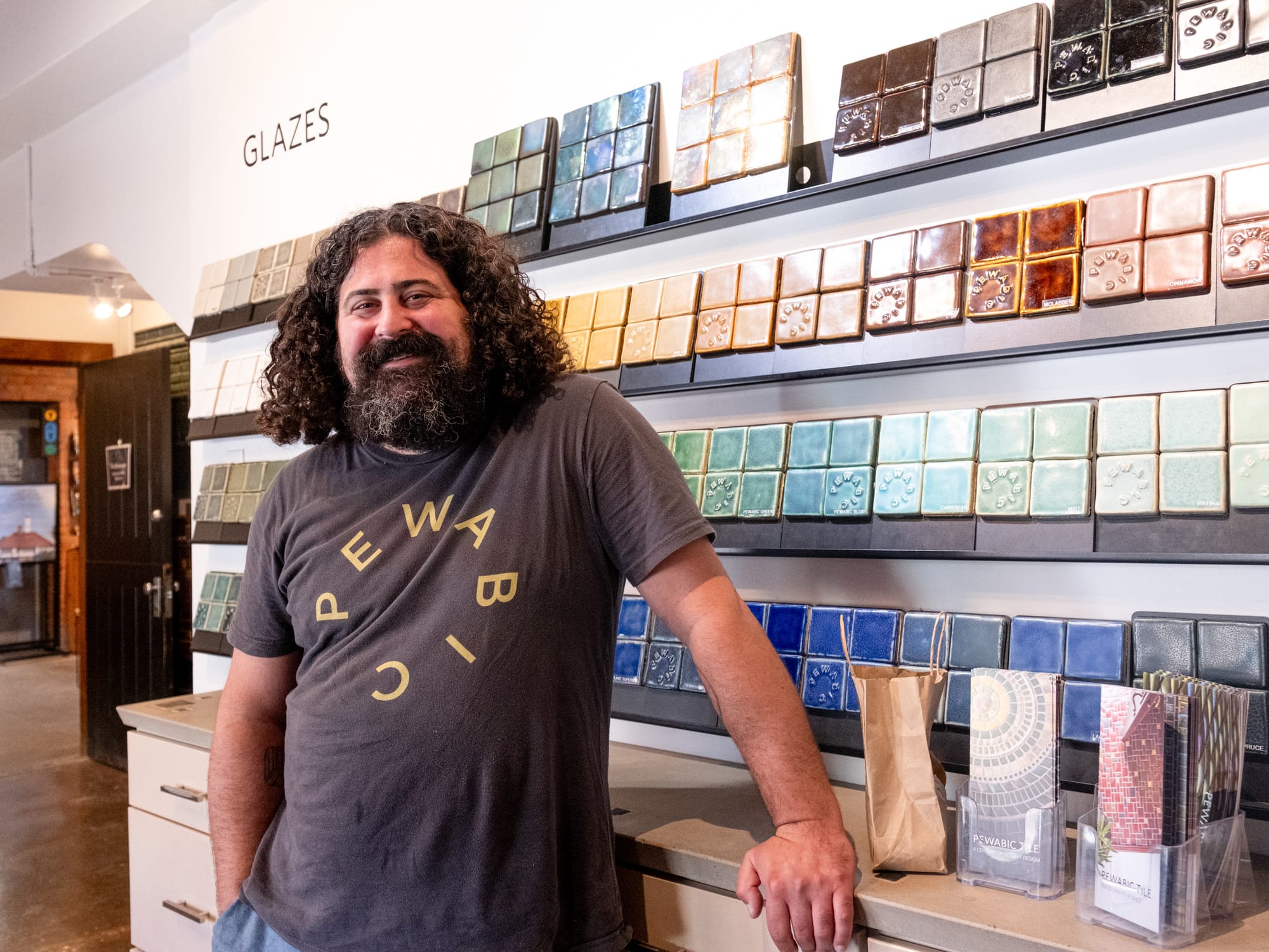 A man stands in front of a white wall with glass tiles displayed on the shelves, wearing a shirt that says ‘Pewabic.