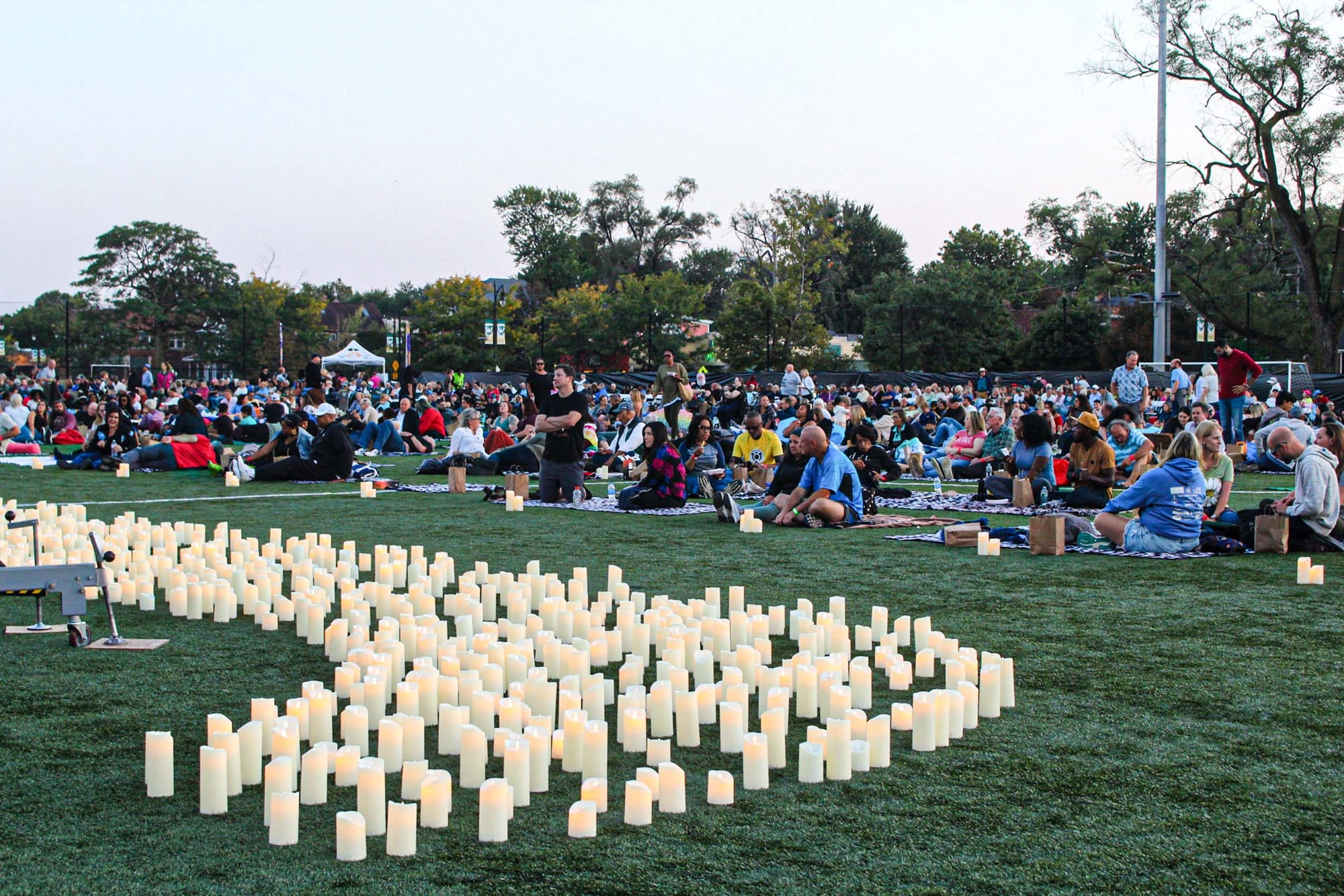 A large crowd of people gathers with blankets and candles facing a candlelit stage while it is still daytime.
