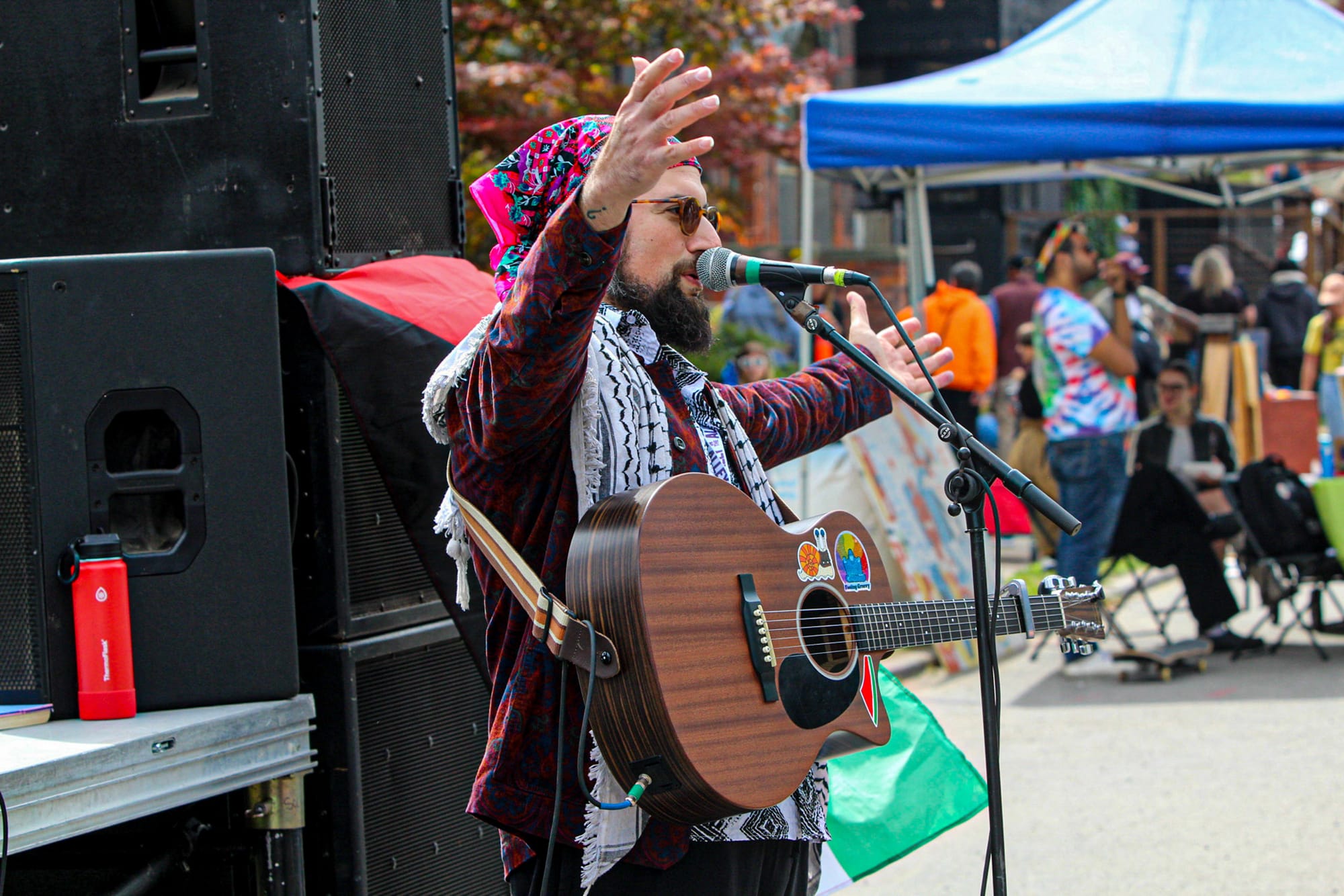 A person with a guitar sings into a microphone with his arms raised at an outdoor event, wearing sunglasses and a Keffiyeh. 