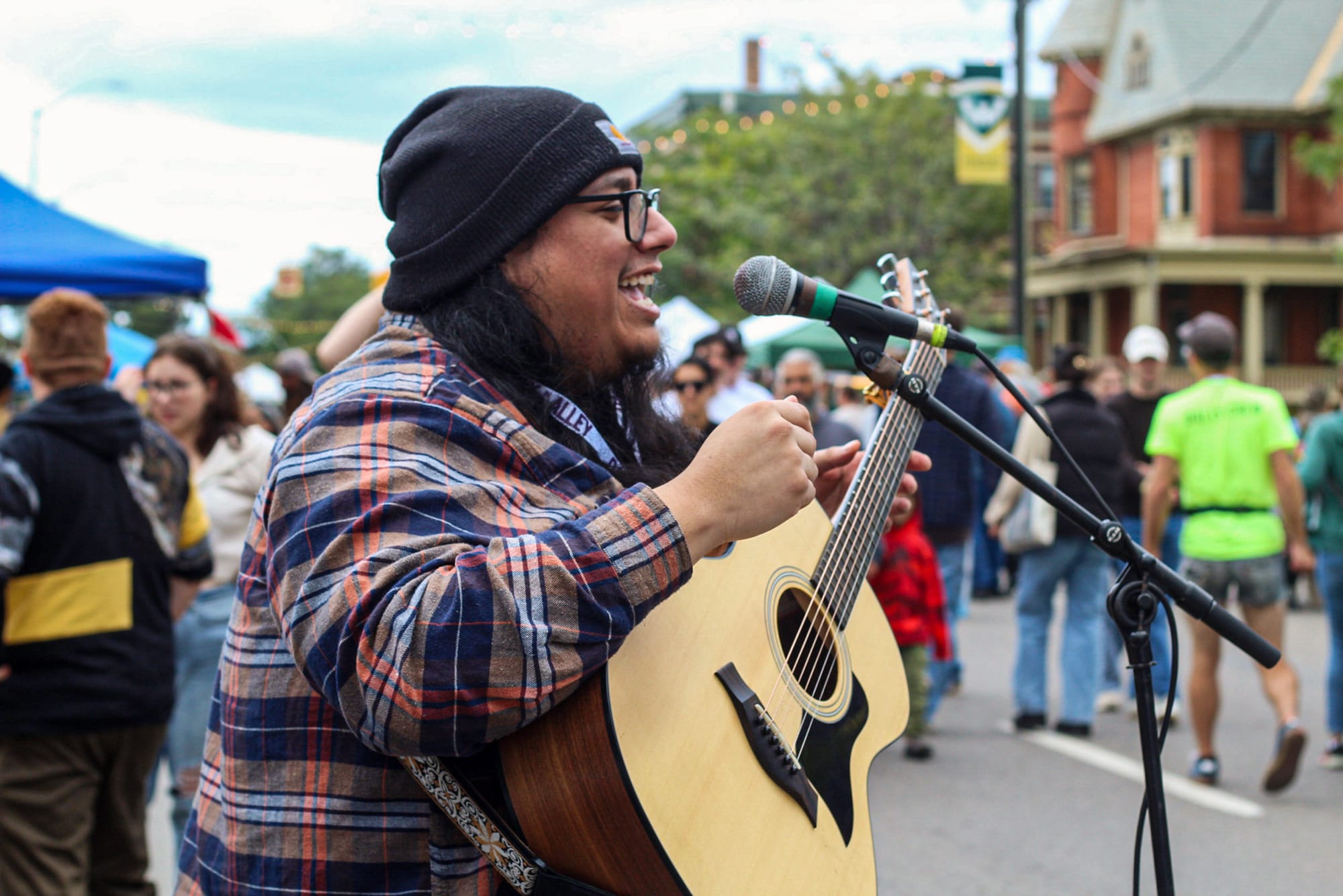 A musician in a flannel and a beanie with a guitar sings into a microphone on the street with a crowd of people behind him. 