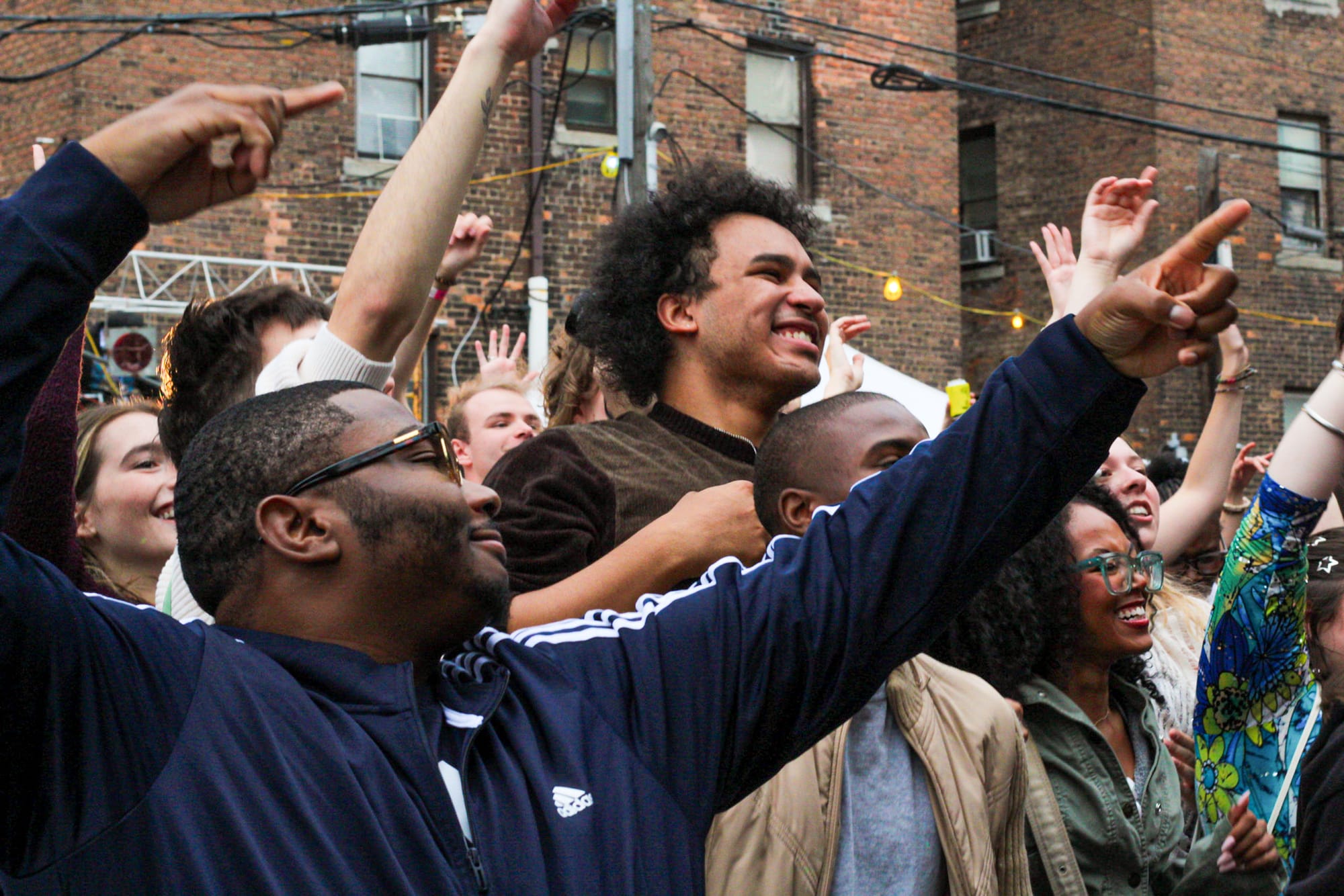 A diverse group of people stand outdoors in front of brick buildings, cheering and raising their arms in excitement.
