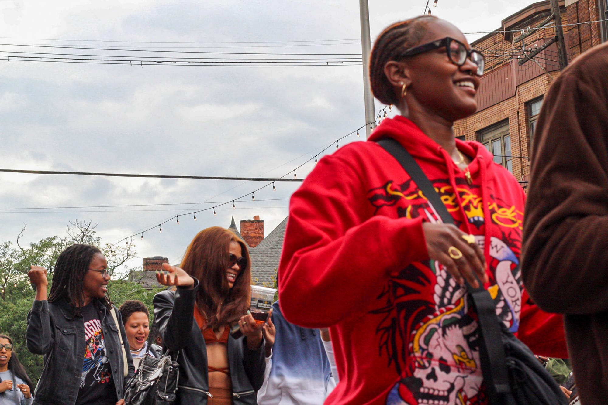 A group of people outdoors smiling and dancing, with one person in a bright red hoodie in the foreground.