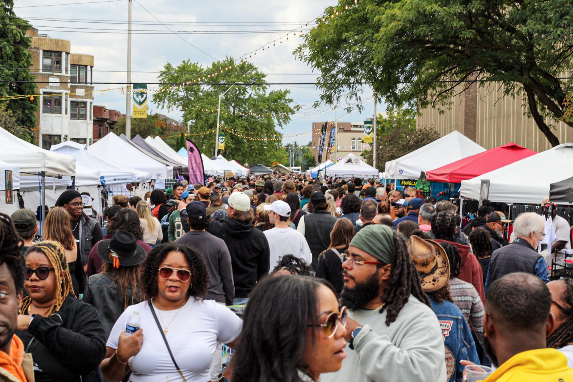 Hundreds of people gathered during the day on a street for a festival with a line of tents on both sides.