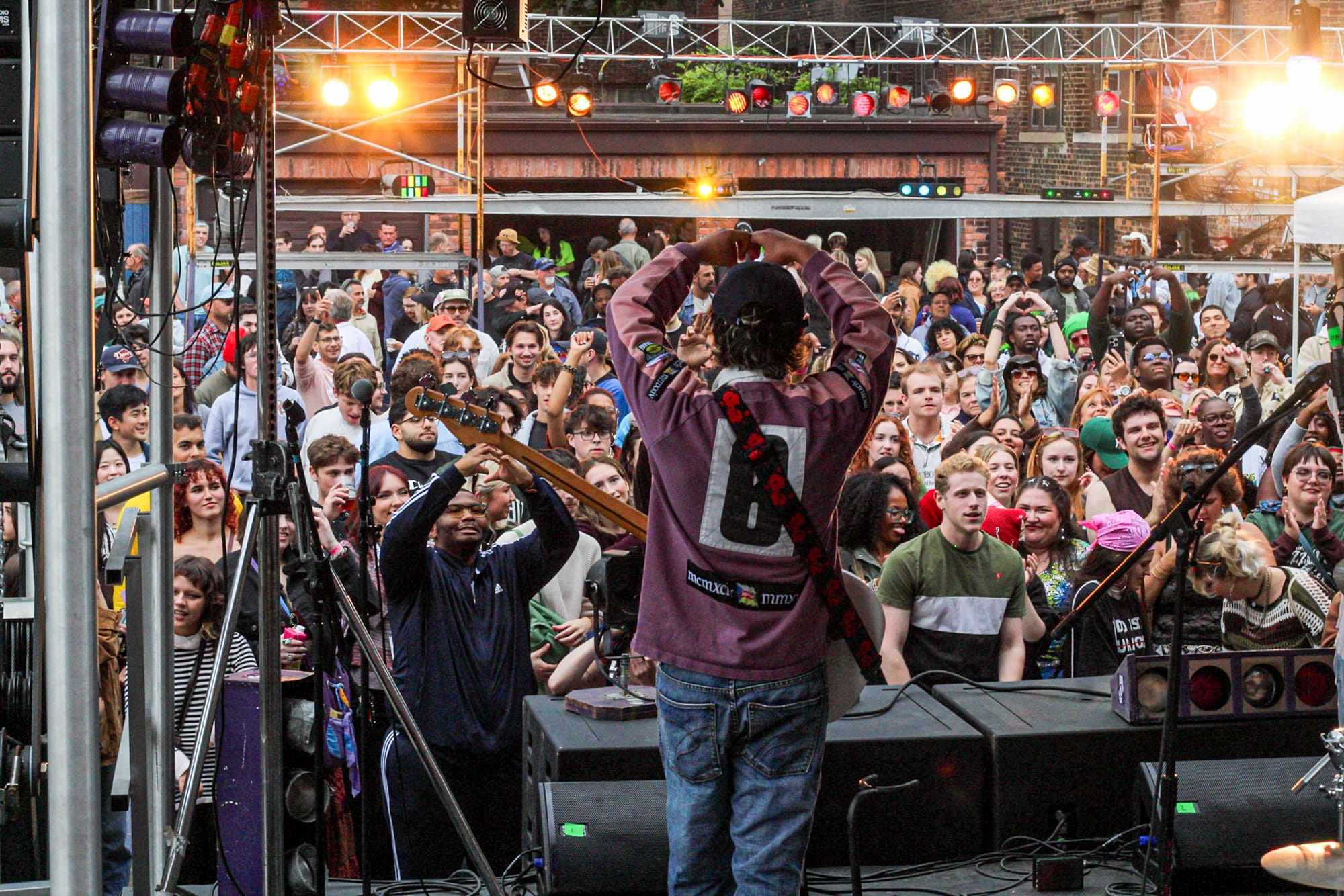 A performer on stage faces a large crowd at an outdoor concert, holding their hands as hearts symbols.
