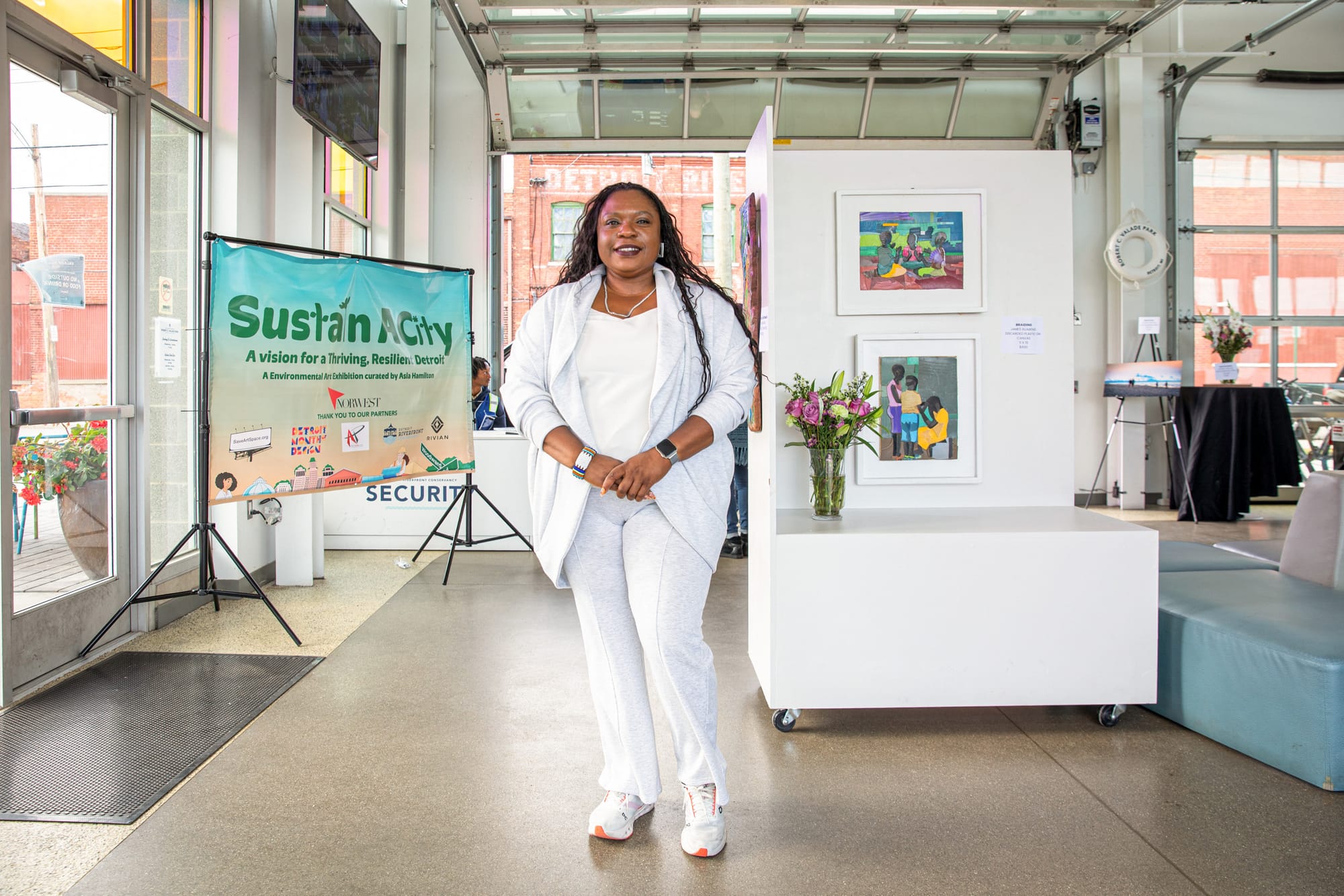 A woman dressed in a white outfit stands smiling behind her, a banner reads “Sustain A City” alongside framed artworks.