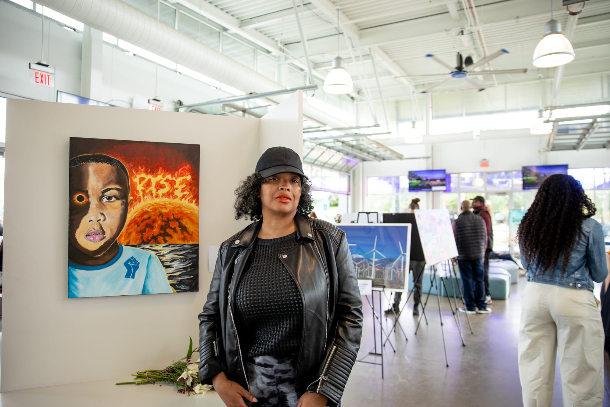 A woman wearing a baseball cap and all black stand in front of an abstracted painting of a young boy.