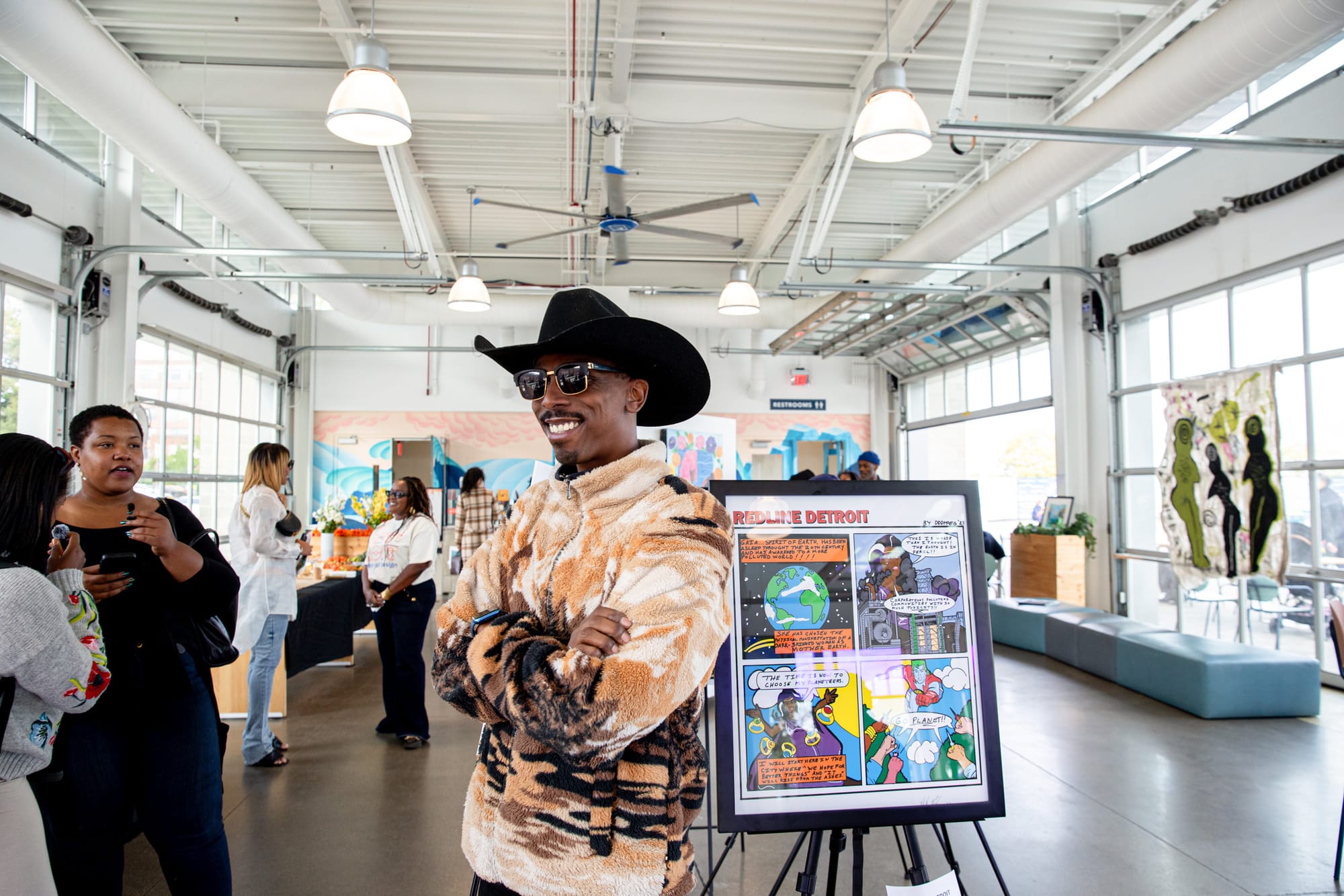 A man in a black cowboy hat stands by an artwork on an easel surrounded by others in a gallery space.