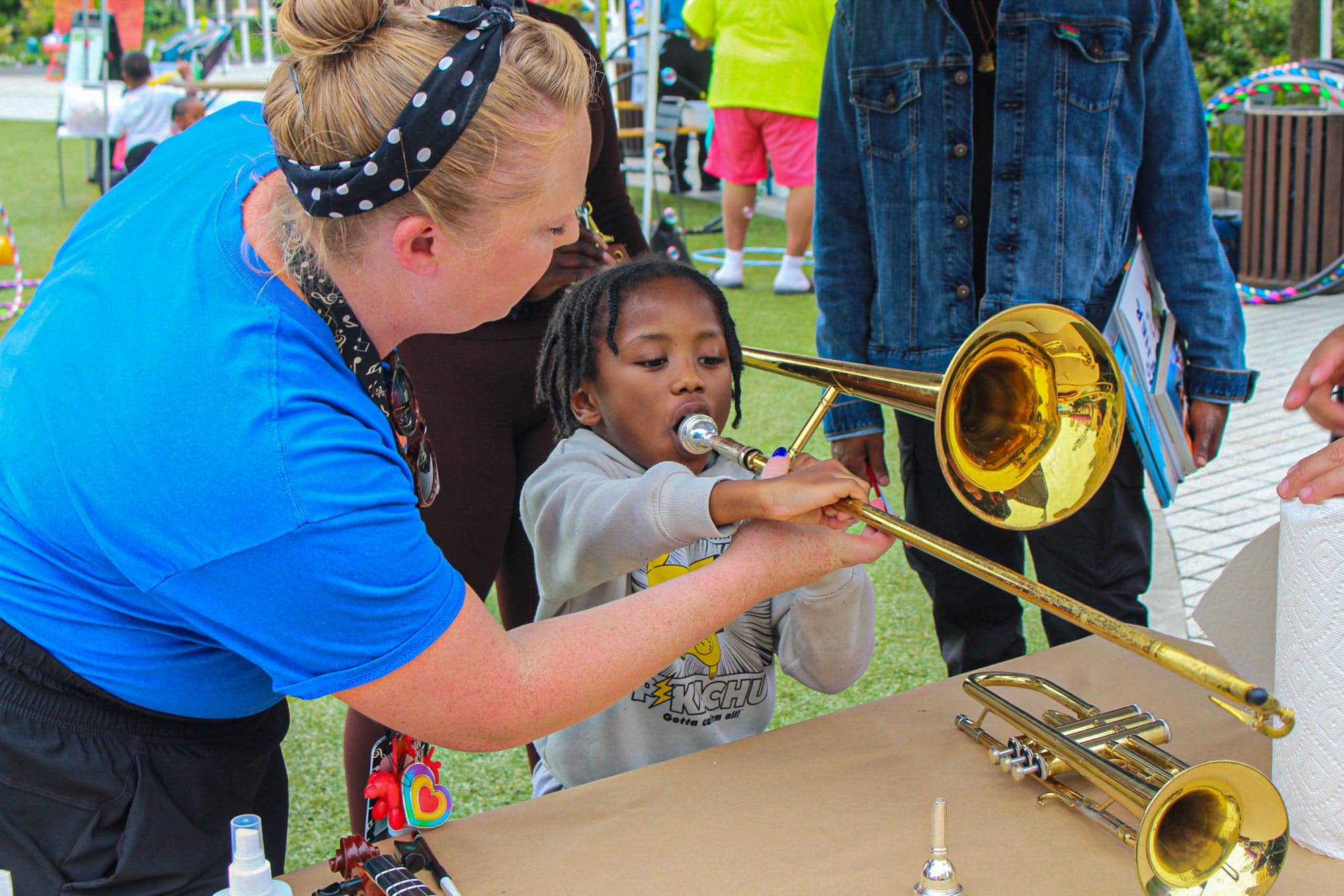 A woman helps a young child play a brass trombone at an outdoor event, with others nearby.