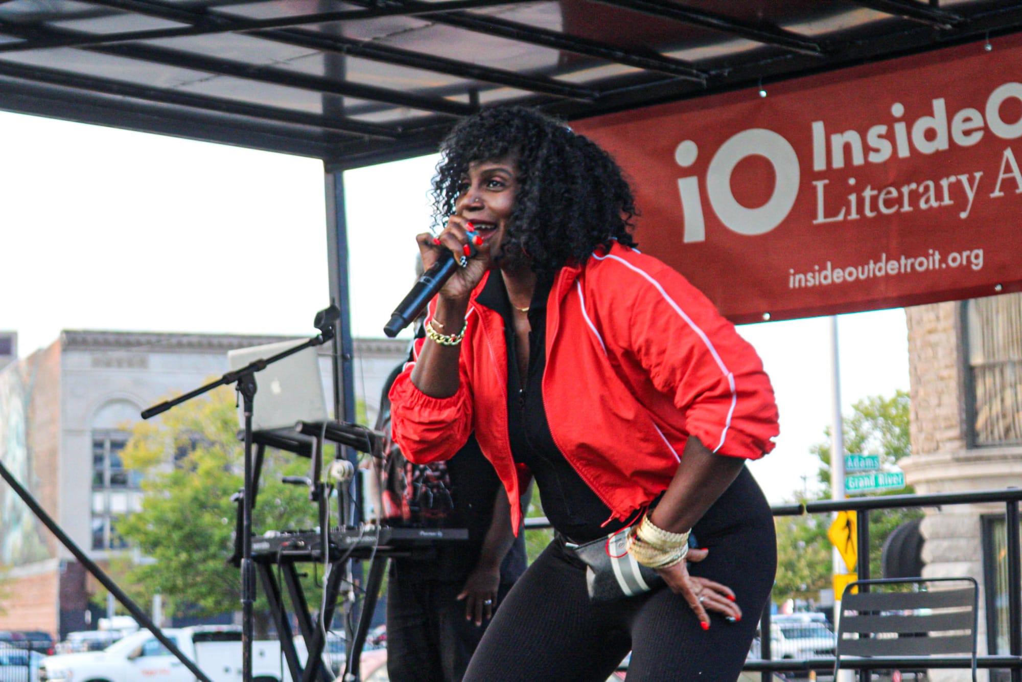 A woman in a red jacket performs on stage with a microphone in one hand and her other hand on her hip as she leans towards the crowd.