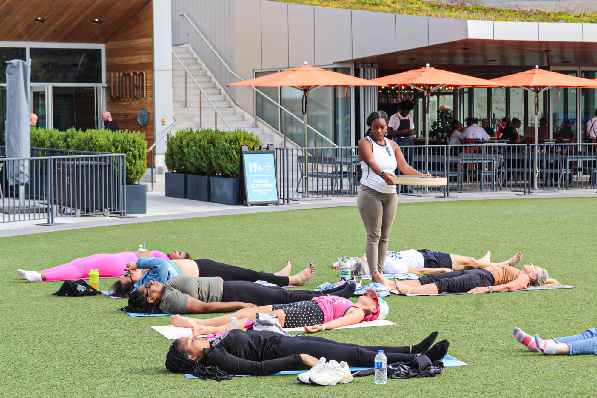 A group of people lying on yoga mats participating in a guided session while a woman stands over them with a wooden instrument.