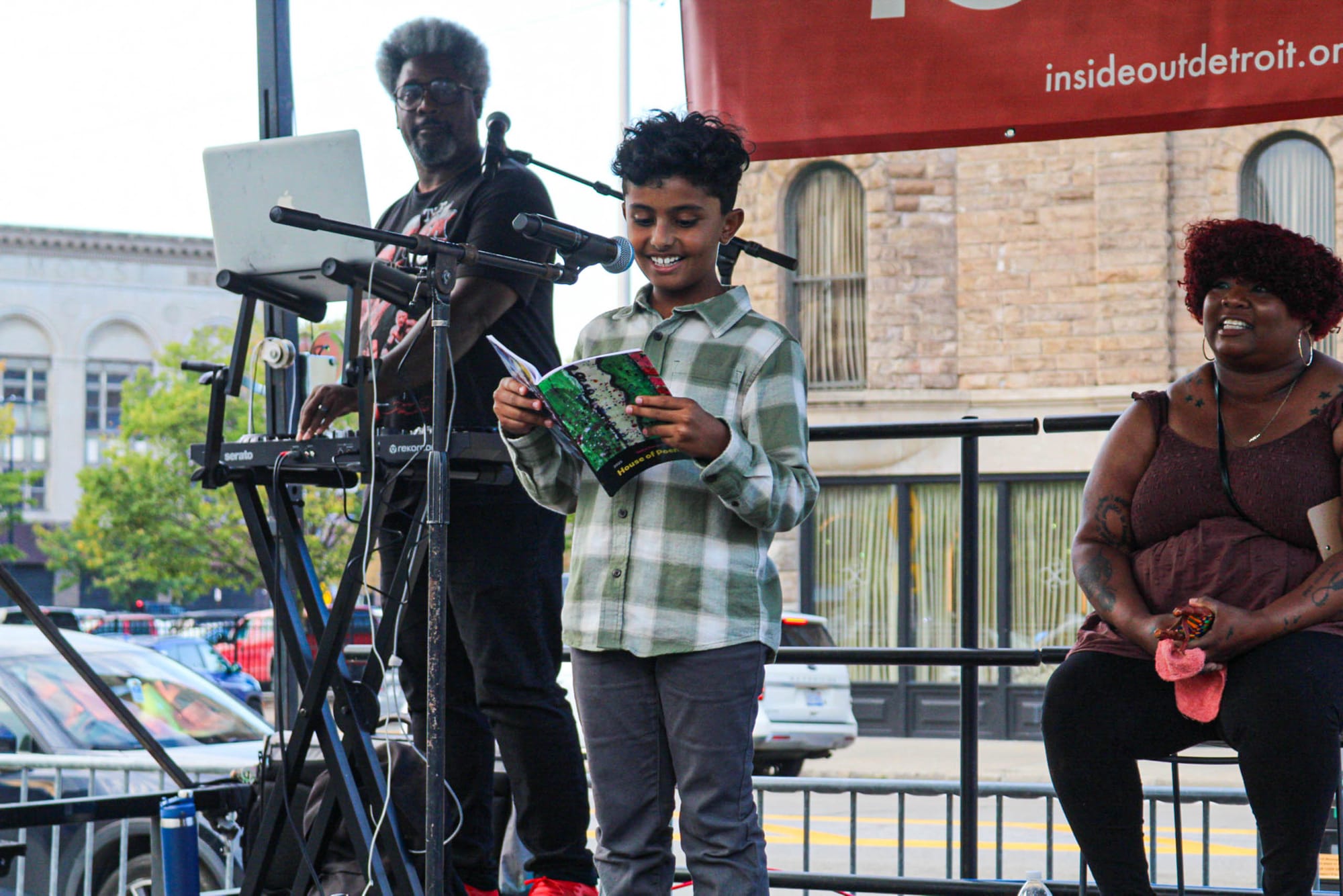 A child is on stage with two adults behind him as he reads a poem from his notebook into a microphone.